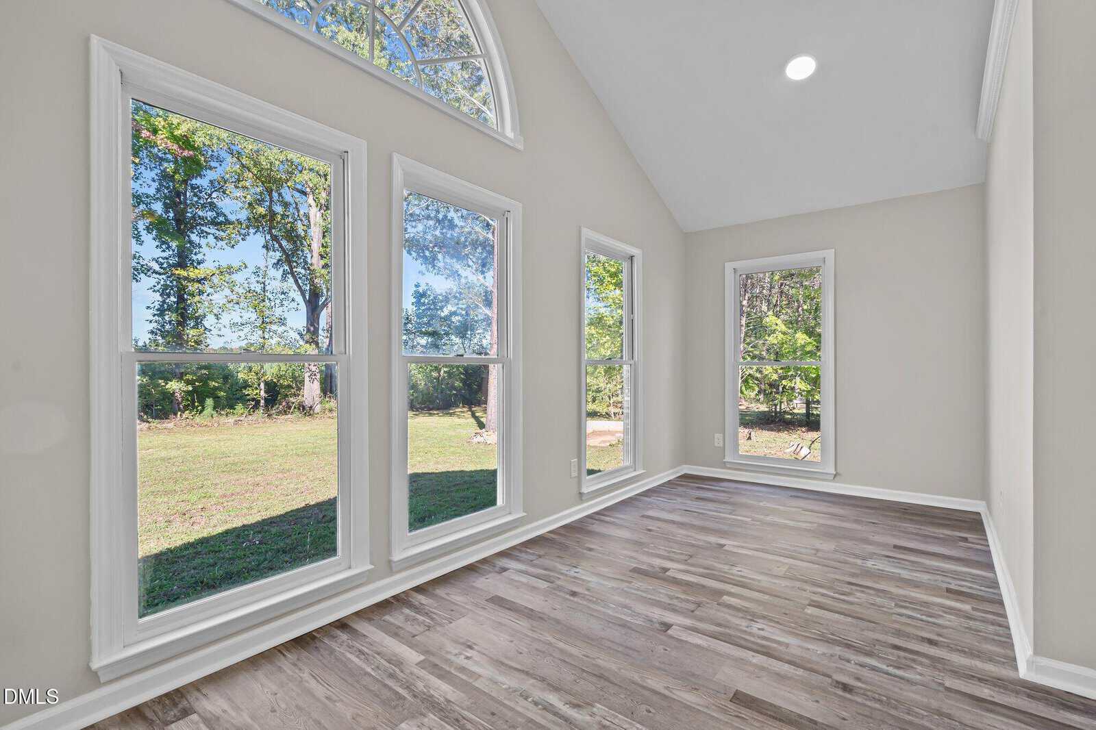 A bright room with large windows, light-colored walls, and wood-look flooring; the windows provide a view of trees and a grassy yard outside. The room has a vaulted ceiling with a circular window at the top.