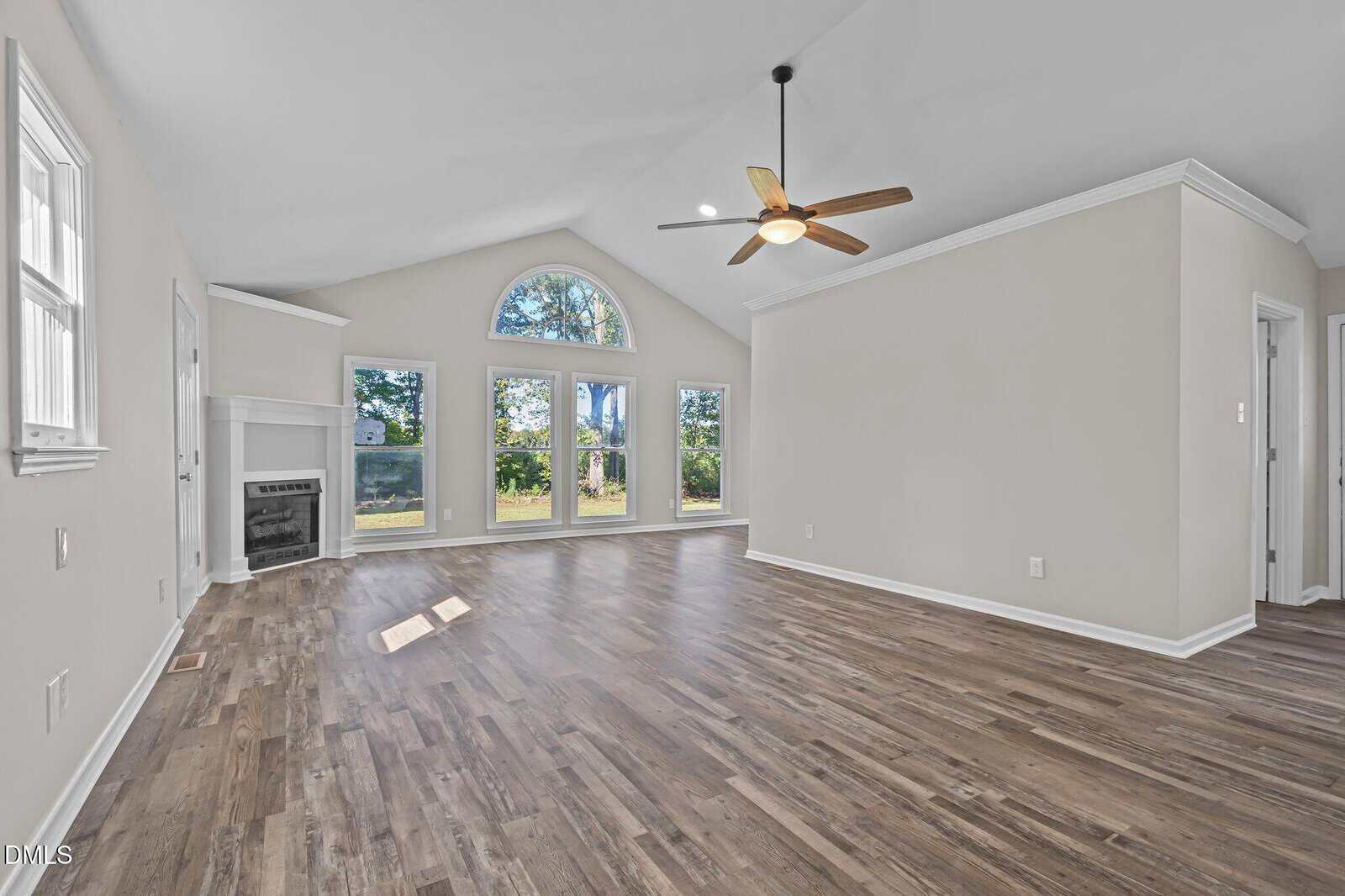 Spacious living room with vaulted ceiling, large arched window, several rectangular windows, ceiling fan, light gray walls, wood-look flooring, and a white fireplace in the corner.