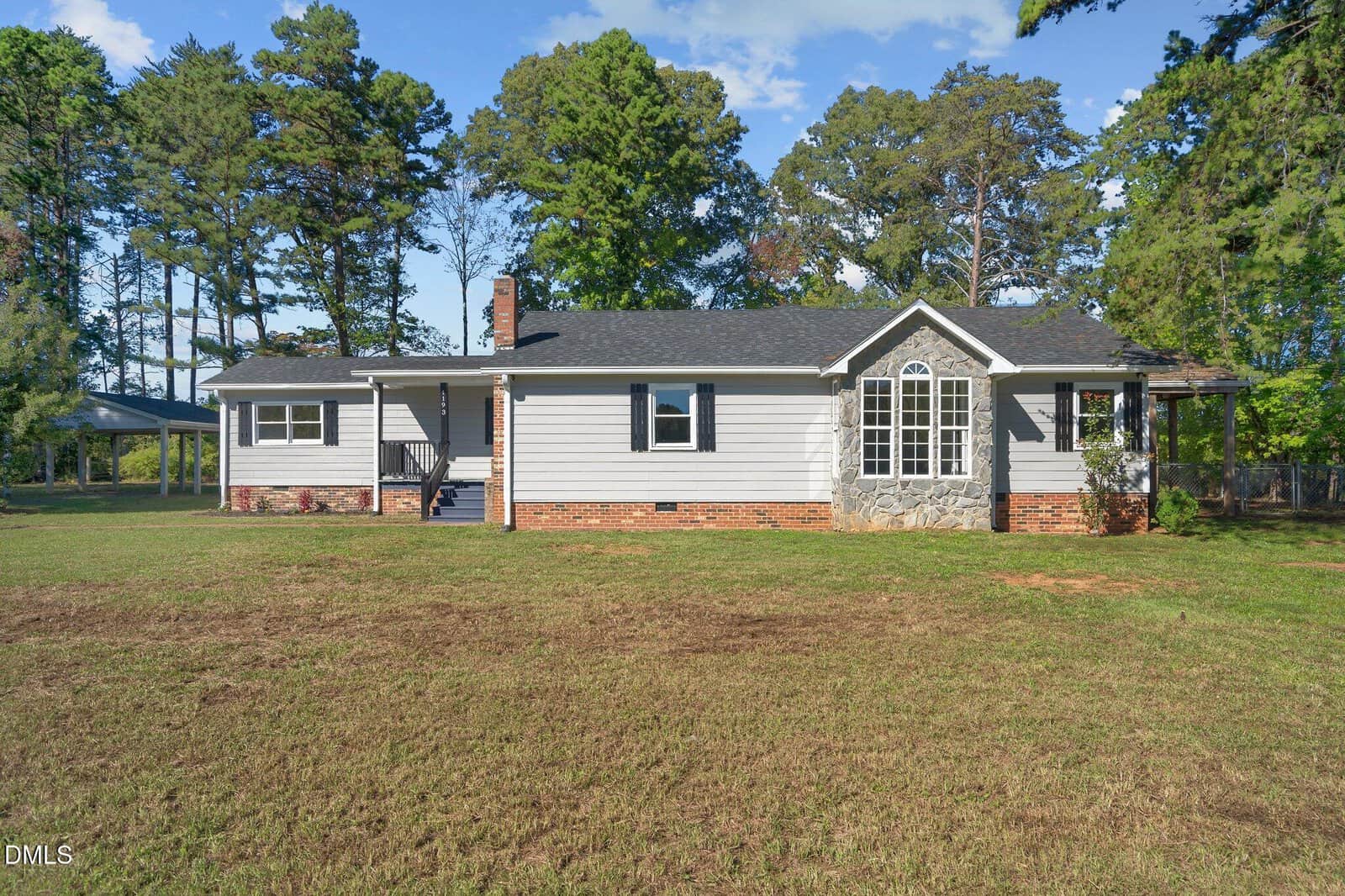 A single-story house with gray siding, black roof, and brick foundation, featuring a prominent set of windows and a small porch. The yard is grassy with sparse vegetation, and tall trees surround the property.