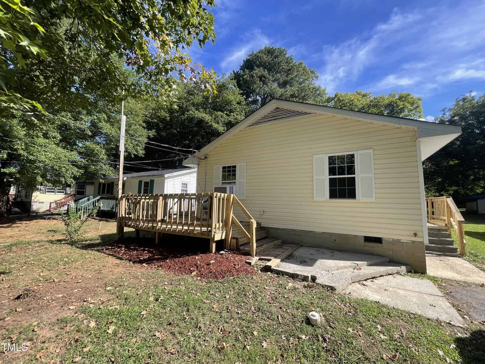 A single-story beige house with white trim and shutters, featuring a wooden accessibility ramp leading to the front door. The yard has grass, mulch, and trees, and it's a sunny day with a blue sky.