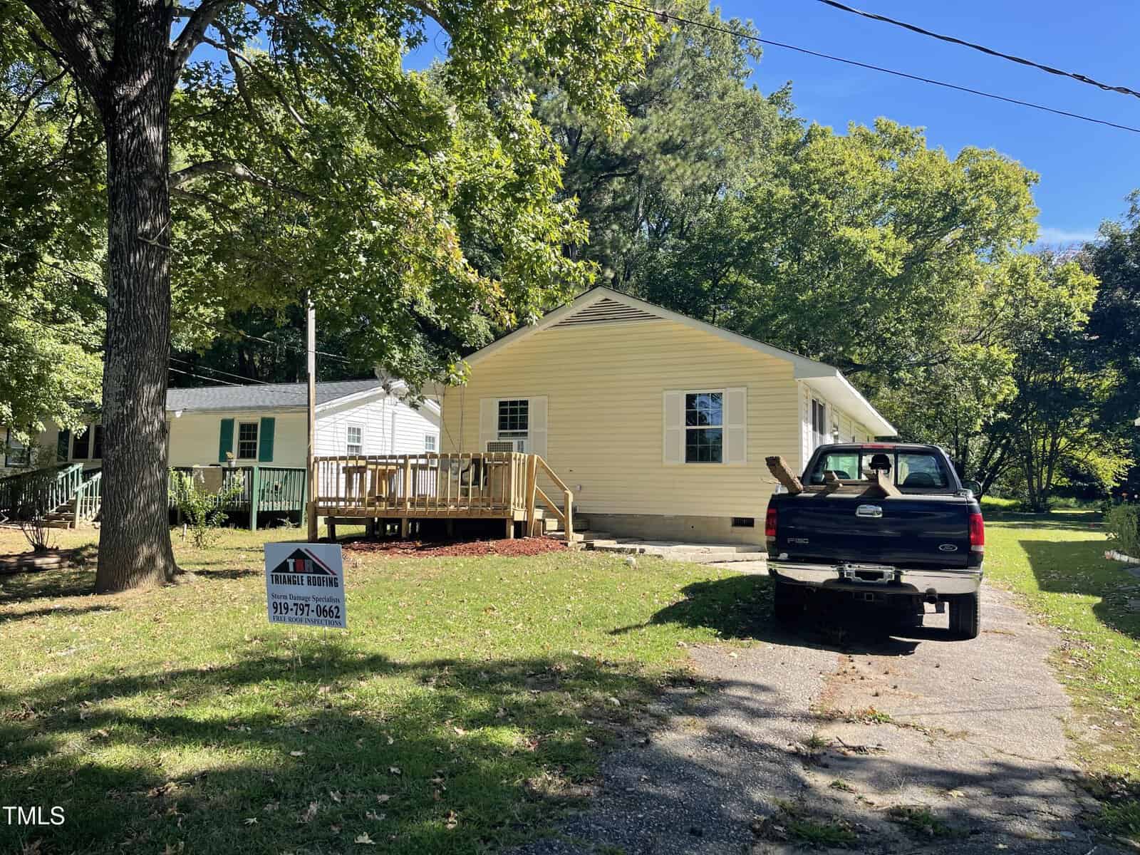 A light yellow single-story house with a wooden front deck sits beside a white house. A black pickup truck is parked in the driveway. A yard sign for a real estate agency is posted on the lawn under a large tree.