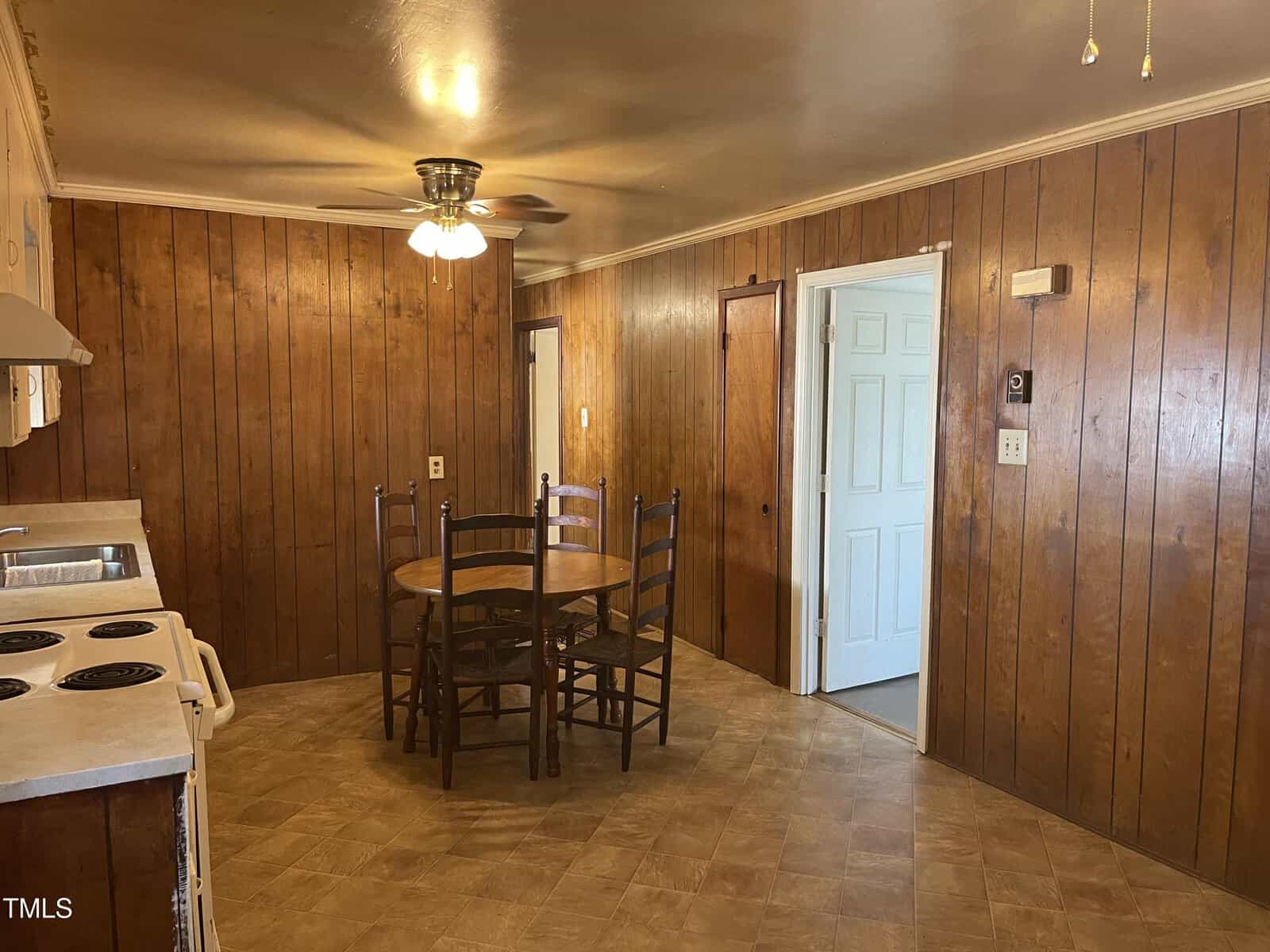 Kitchen with wood-paneled walls, a ceiling fan with lights, a round wooden table with four chairs, white stove and counter, and a door leading to another room. The floor has a tan tile pattern.