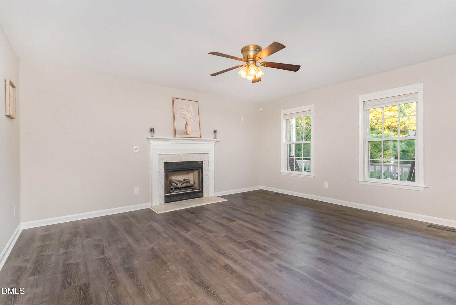A spacious living room with light beige walls, dark wood flooring, a white fireplace with art above it, a ceiling fan with lights, and two large windows letting in natural light.