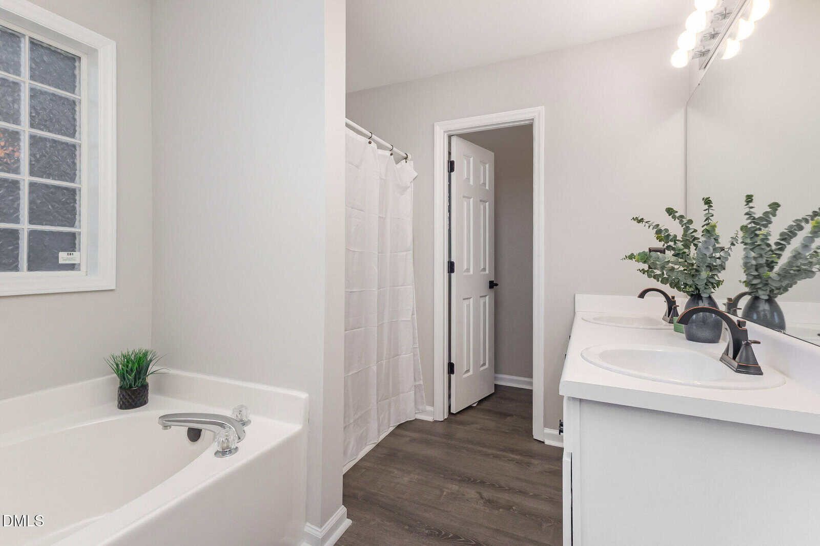 A bathroom with a white bathtub, a double sink vanity with two mirrors, a vase with greenery, a shower with a curtain, and a door. The walls are light-colored and the floor has dark wood-style flooring.