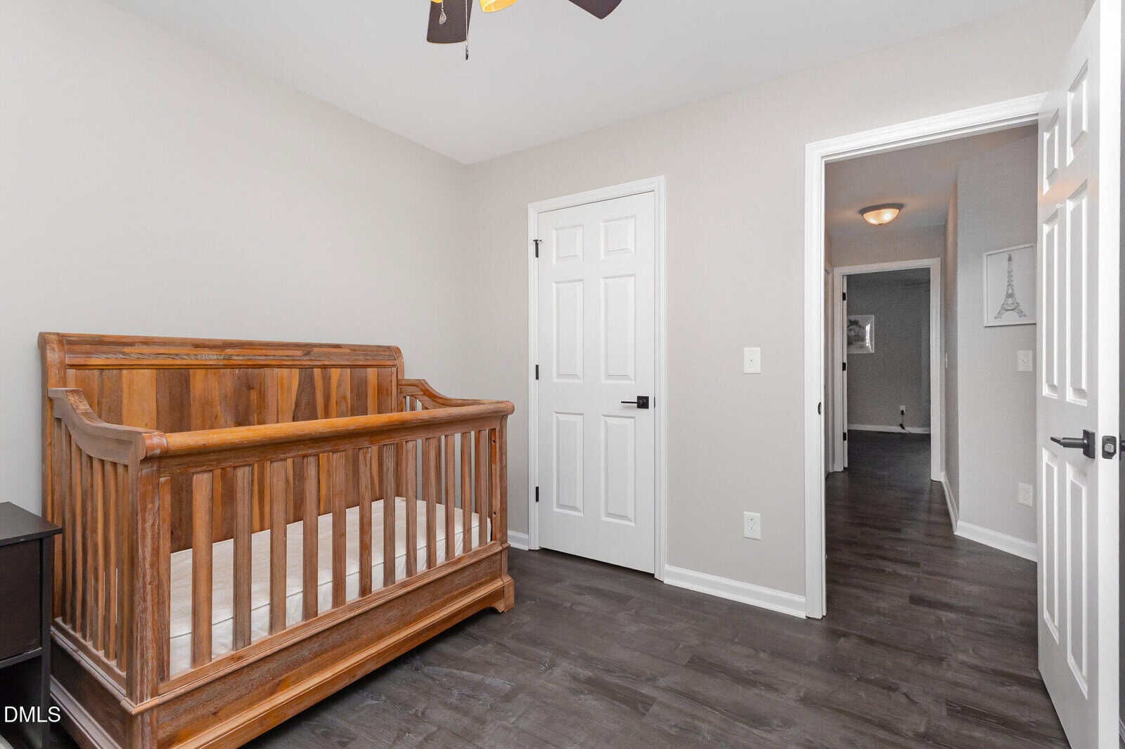 A nursery room with a wooden crib, white walls, dark wood flooring, and two white doors; one door is open, revealing a hallway with a light fixture and framed artwork on the wall.