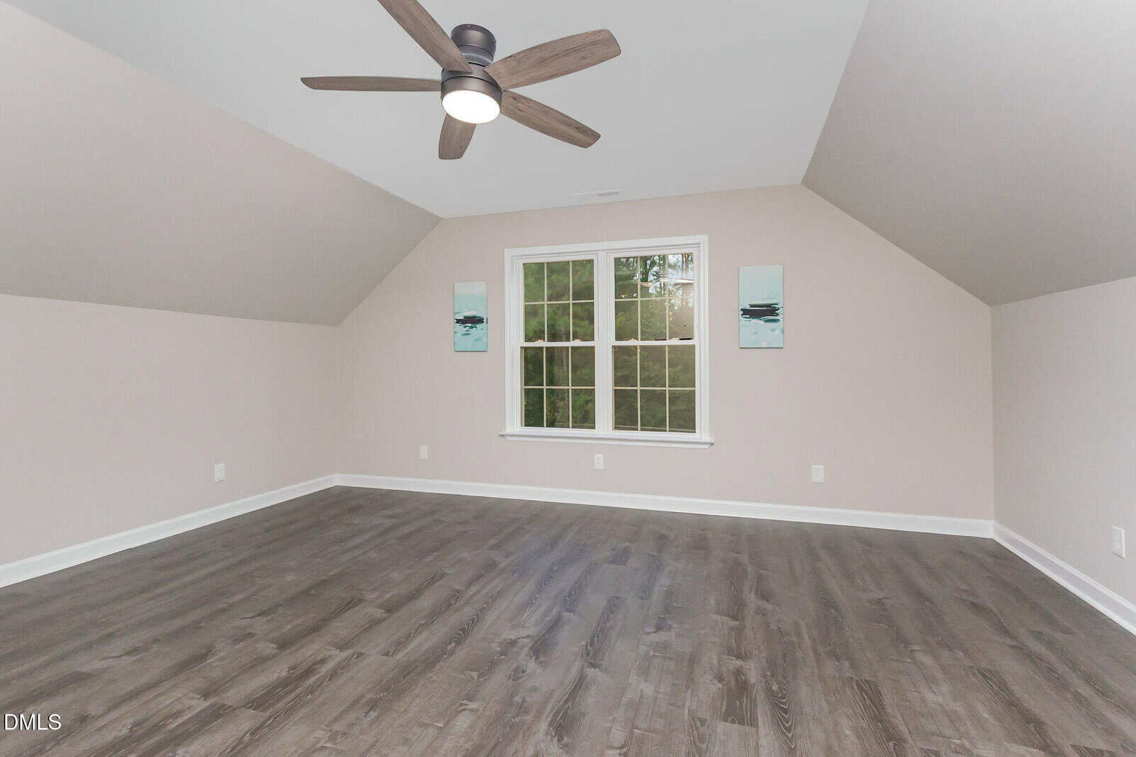 An empty room with light-colored walls, a window centered on the far wall, two small pieces of abstract art, dark wood flooring, and a ceiling fan with wooden blades.