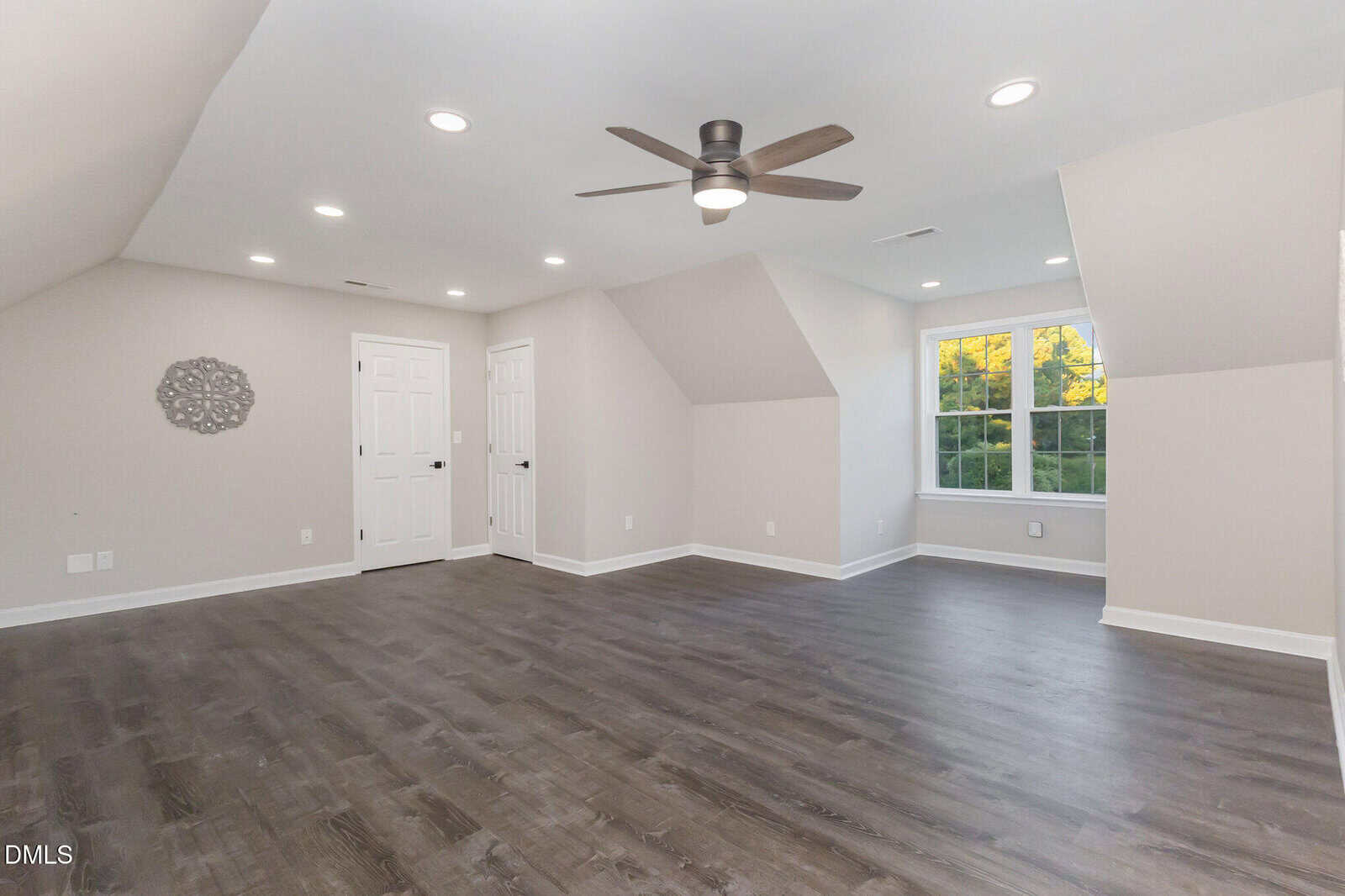 Spacious, empty room with light walls, dark wood floors, ceiling fan, recessed lighting, two white doors, and a large window letting in natural light.