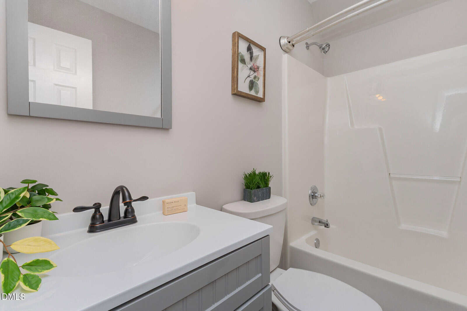 A modern bathroom with a white sink, gray vanity, black faucet, mirror, potted plants, wall art, and a white bathtub-shower combo with a shower curtain rod.