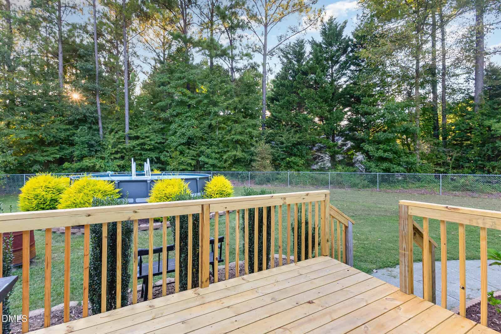 A wooden deck overlooks a grassy backyard with a chain-link fence, green trees, and an above-ground pool in the background. The scene is bright and peaceful.