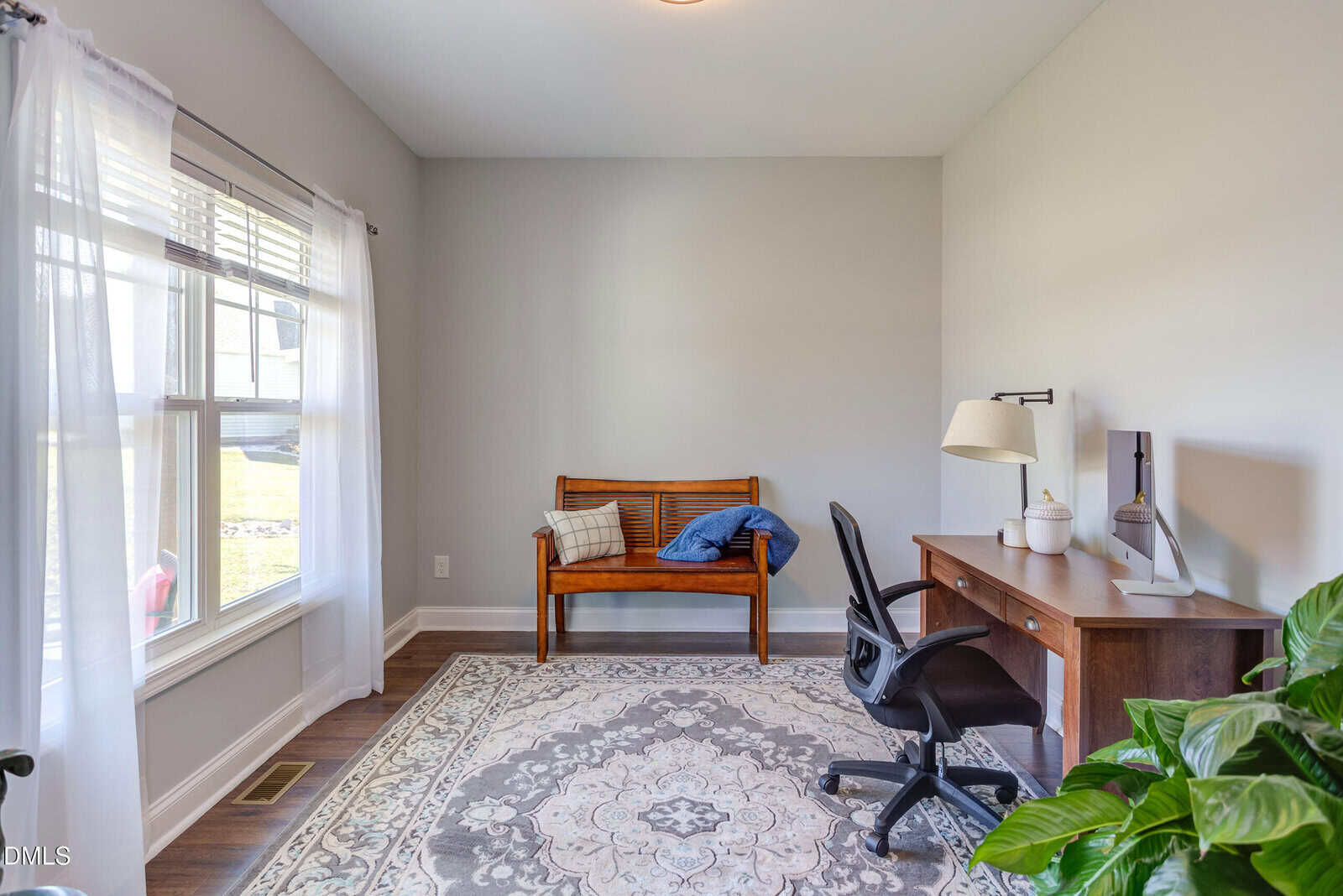 A bright home office with a large window, wooden desk, black chair, bench with cushions, light gray walls, patterned rug, and a potted green plant. Natural light fills the tidy, minimalist space.