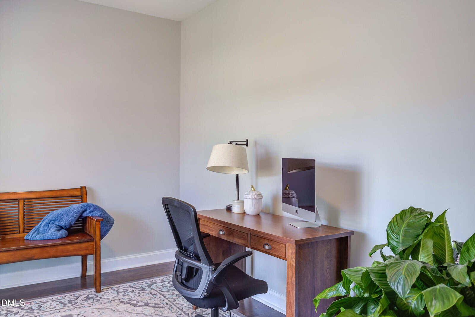 A home office with a wooden desk, black office chair, desktop computer, table lamp, and decorative jars. A plant sits on the floor nearby, and a wooden bench with a blue throw is against the wall.