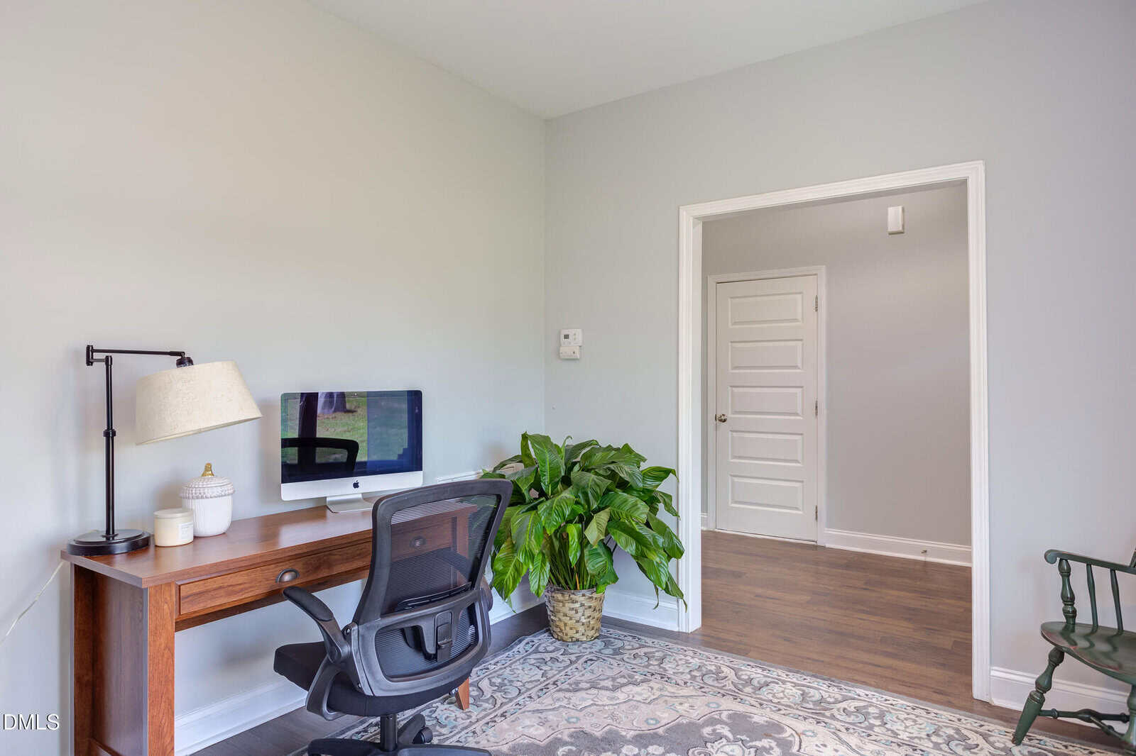 A home office with a wooden desk, computer, lamp, chair, potted plant, and a decorative rug. The room has light-colored walls and an open doorway leading to a hallway with white doors.