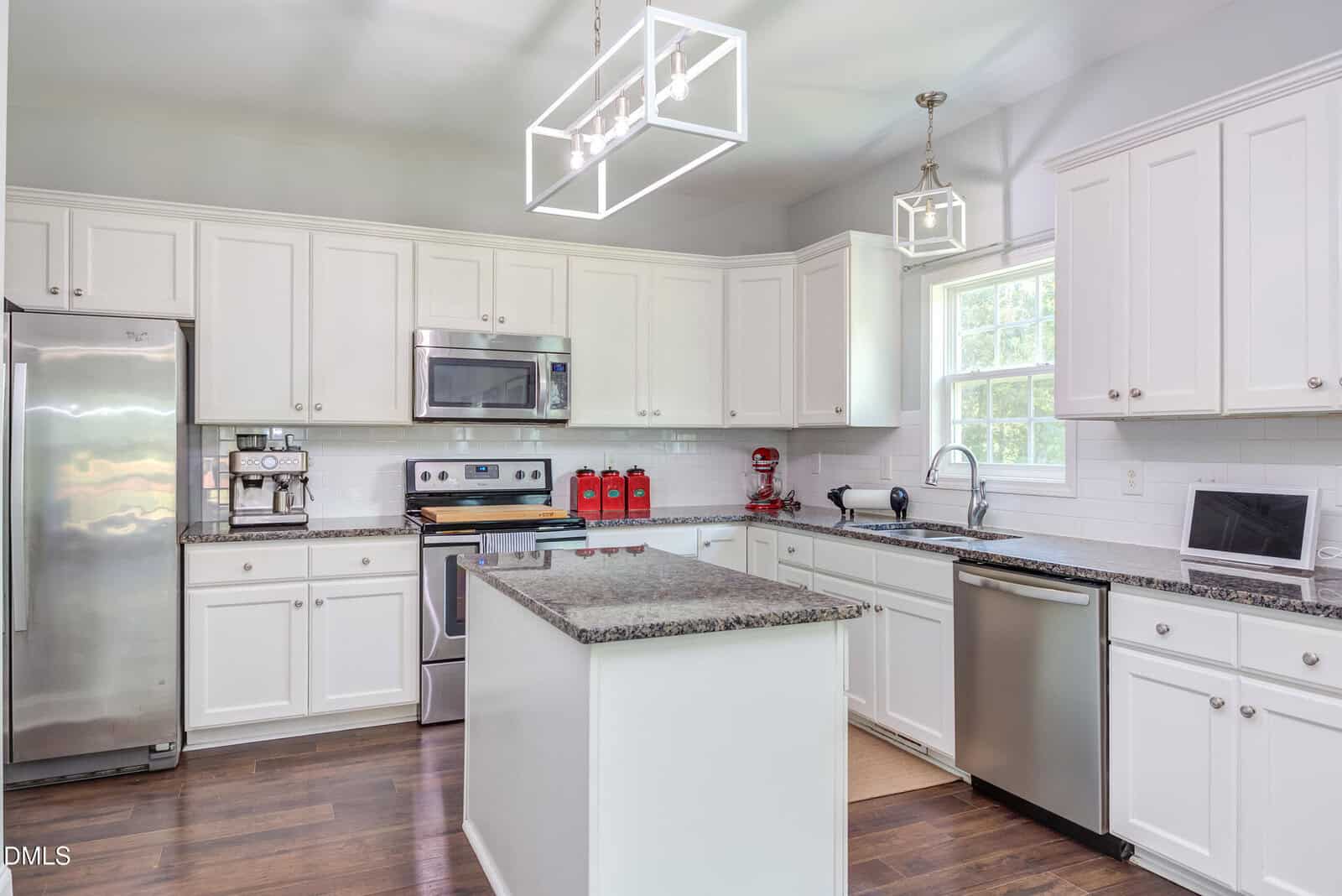 Modern kitchen with white cabinets, stainless steel appliances, granite countertops, an island in the center, and two geometric pendant lights. A window brings in natural light above the sink.