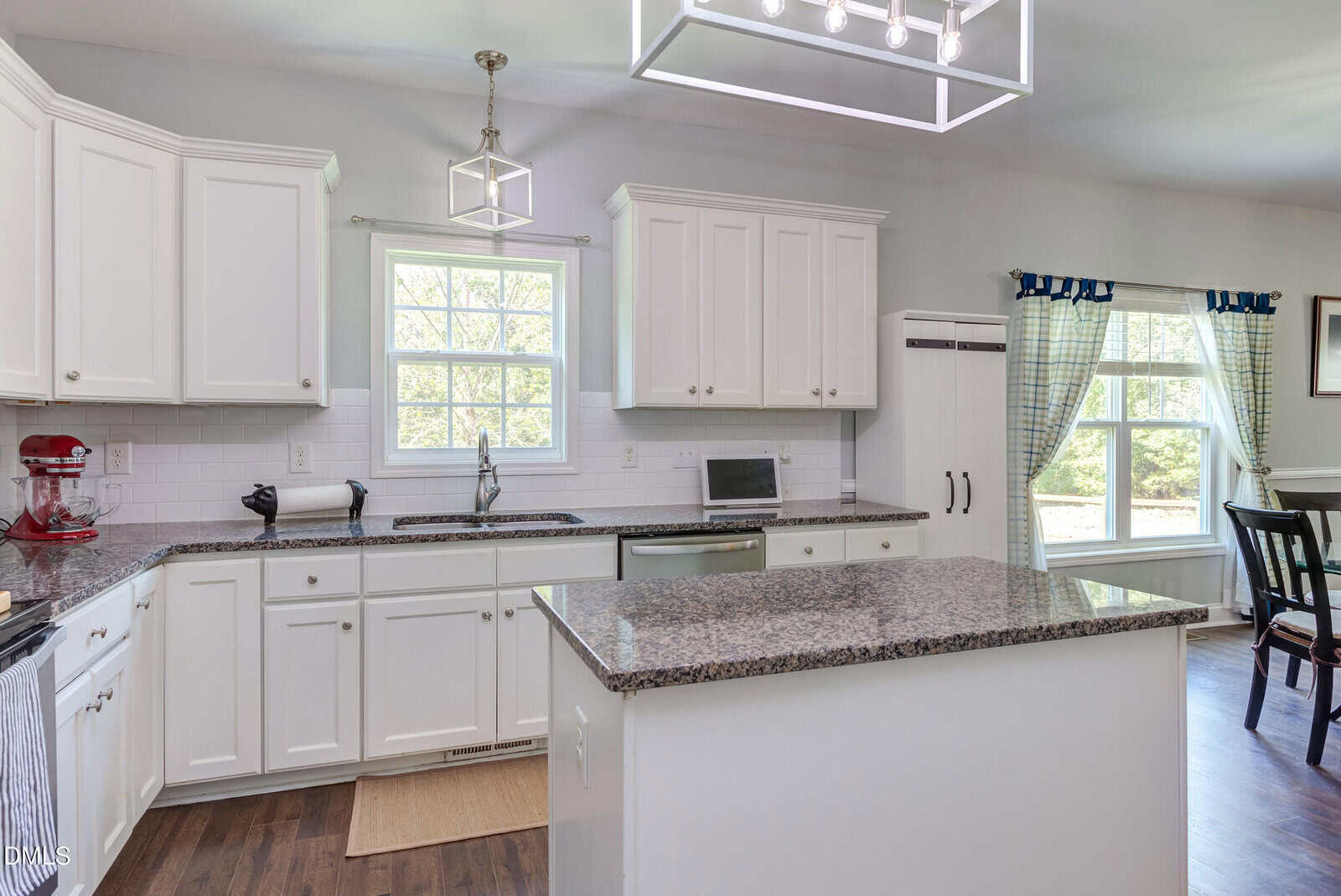 Modern kitchen with white cabinets, granite countertops, an island, stainless steel appliances, a stand mixer, and large windows letting in natural light. Dining table visible in the background.