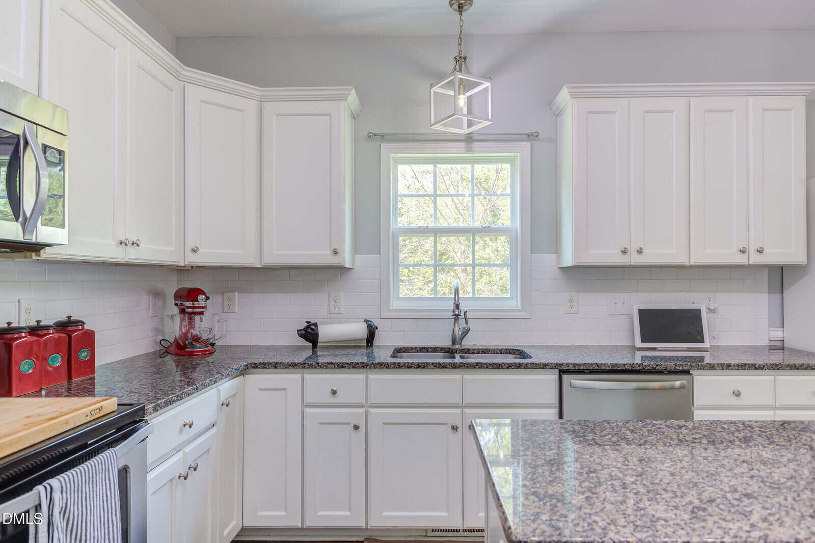 Bright kitchen with white cabinets, granite countertops, stainless steel appliances, a red mixer, a window above the sink, paper towels, and a small TV or monitor on the counter.