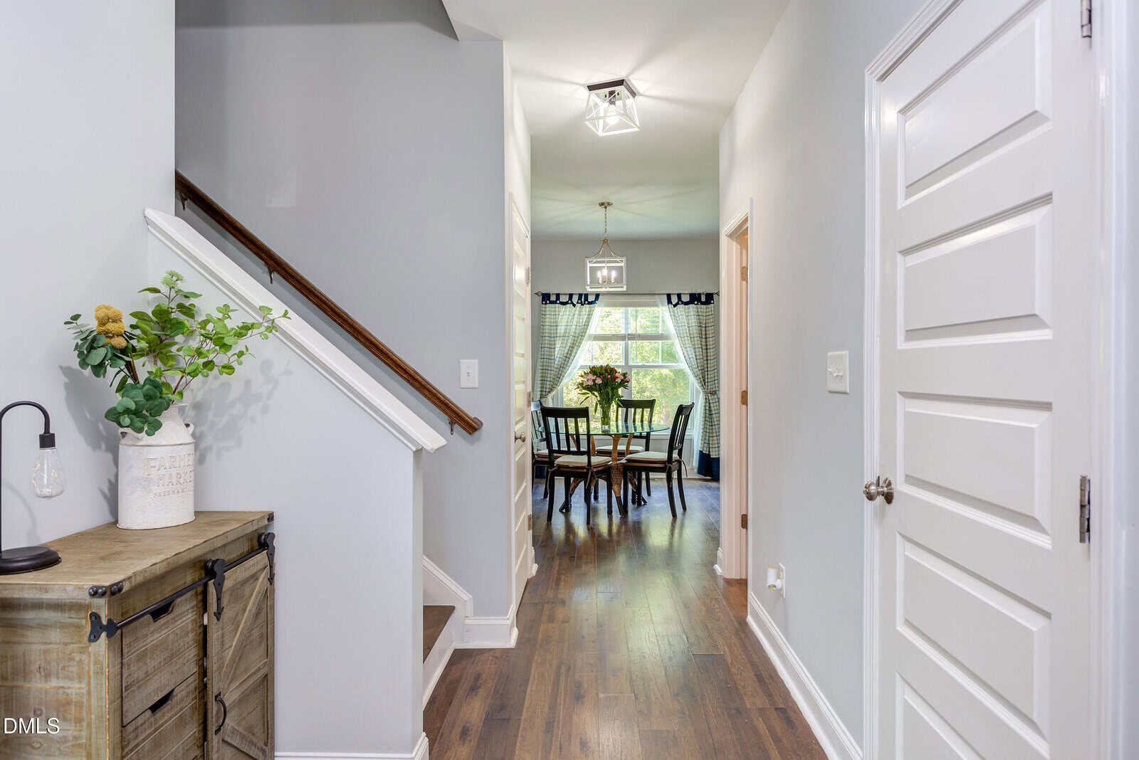 A bright hallway with light gray walls and wood flooring leads to a dining area with large windows. A staircase is on the left, and a chest with a plant and lamp sits beneath it. White doors and a hanging light fixture are visible.