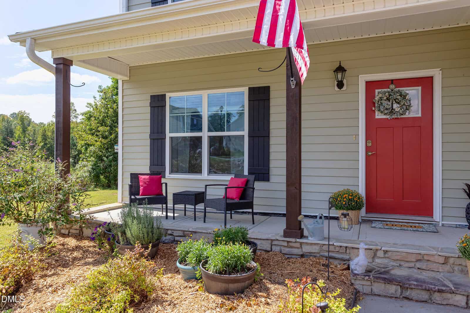 A front porch with two black chairs with red cushions, a small table, potted plants, a red door with a wreath, and an American flag hanging above. The area is landscaped with mulch and stone edging.