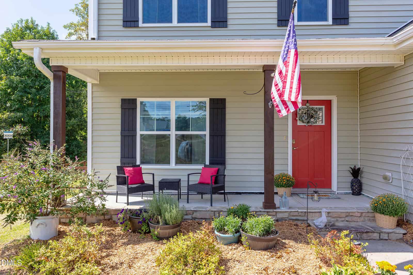 A front porch with two black chairs, red cushions, a small table, potted plants, an American flag, and a red front door on a beige house with black shutters and a garden bed in front.