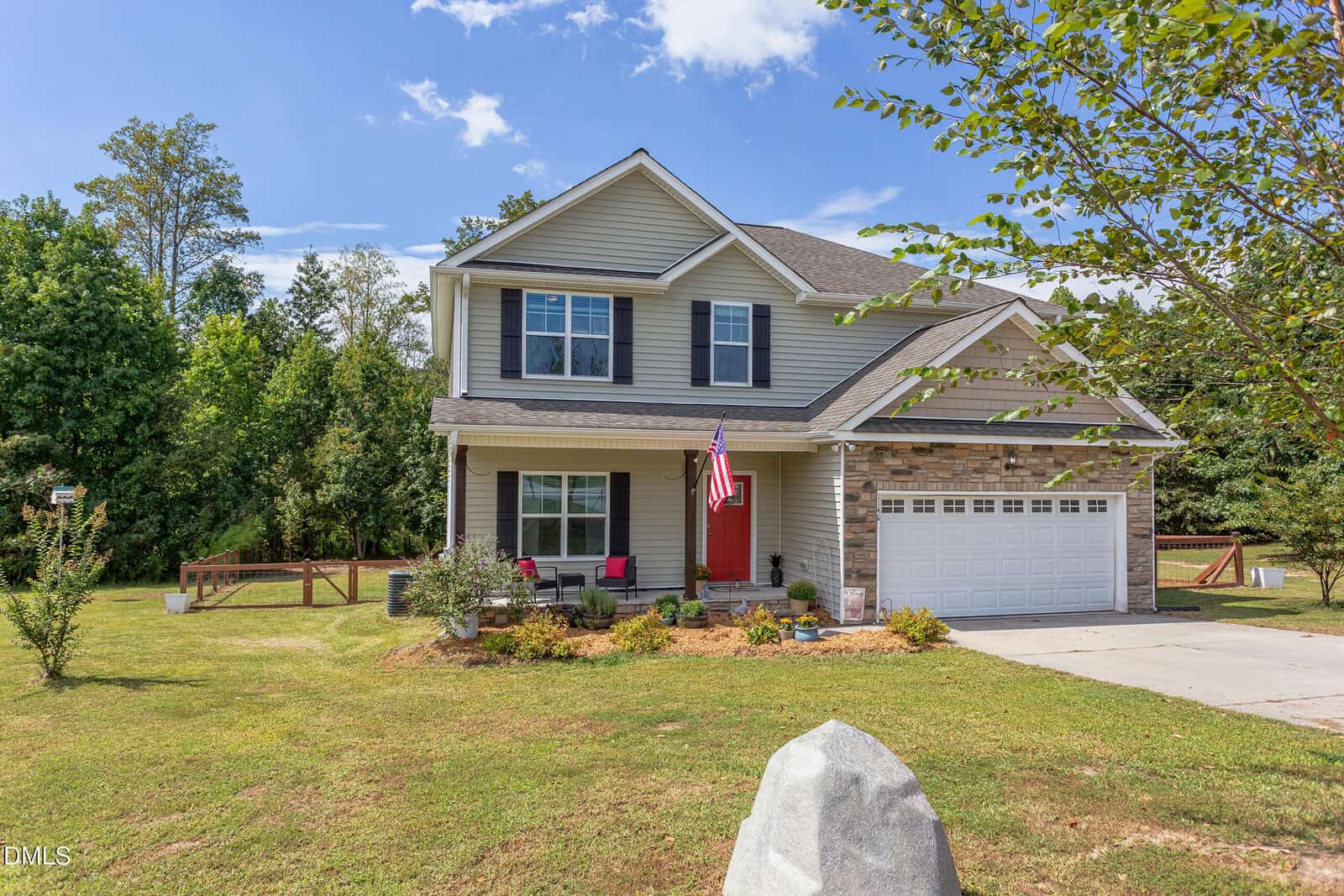 Two-story suburban house with gray siding and brick accents, a front porch with red and pink chairs, an American flag, a two-car garage, and a well-kept lawn bordered by trees under a blue sky.