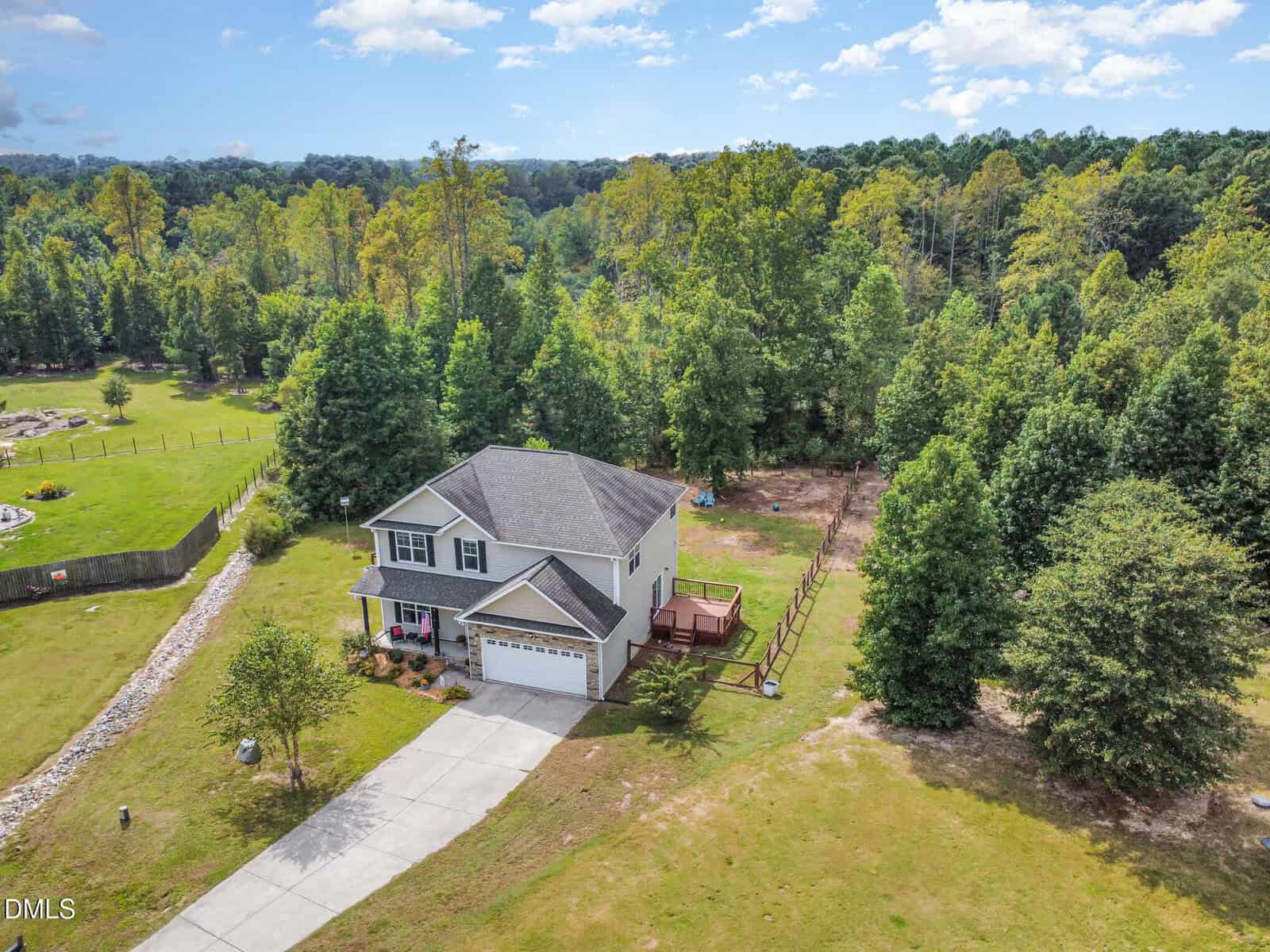 A two-story house with a gray roof and attached garage sits on a spacious, grassy lot bordered by trees. A wooden deck is in the backyard, and a long driveway leads to the house. Dense forest surrounds the property.