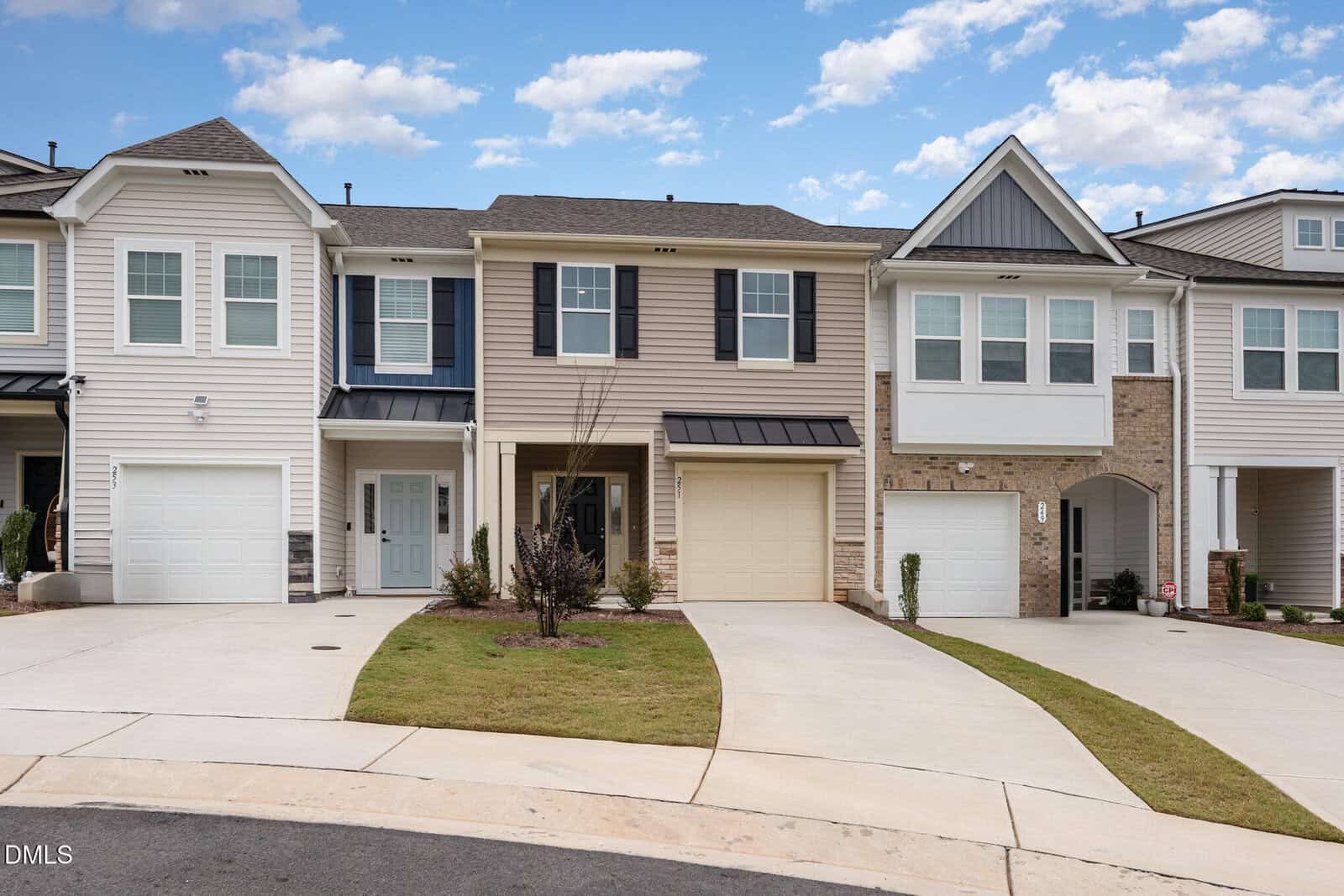 Row of modern two-story townhouses with garages and small front lawns, set along a curved driveway. The homes have beige and white siding, black shutters, and a partly cloudy sky overhead.