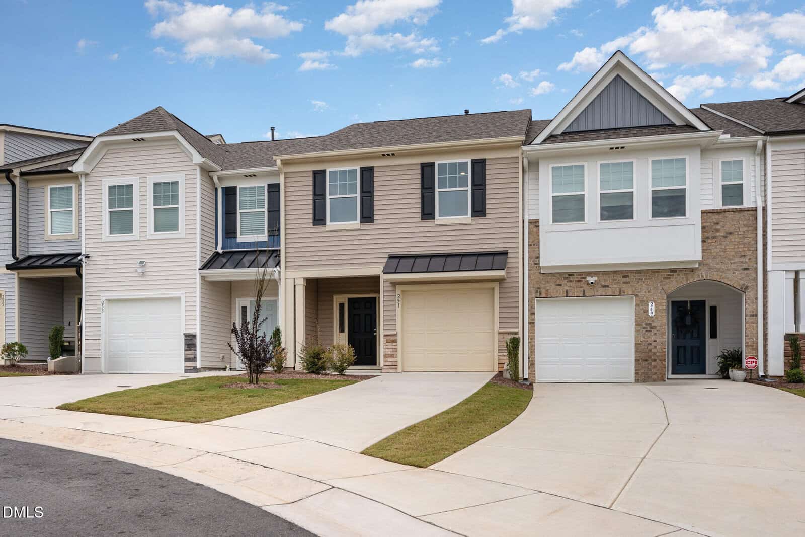Row of modern two-story townhomes with attached garages, beige and white siding, black shutters, and small front lawns along a curved concrete driveway under a partly cloudy sky.