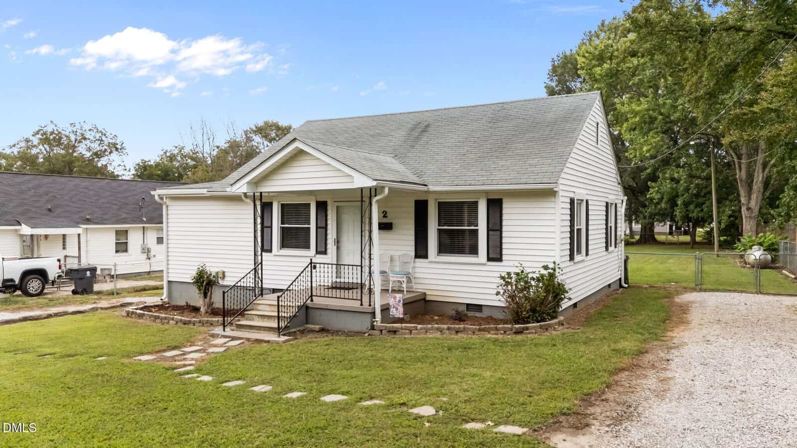 Charming single-story white house with a gray roof, front porch, and black railings, perfectly situated at 2 Dover Street in Franklinton. The yard features a stone walkway bordered by grass, while a gravel driveway runs alongside the house. Mature trees provide a peaceful backdrop to this inviting home.