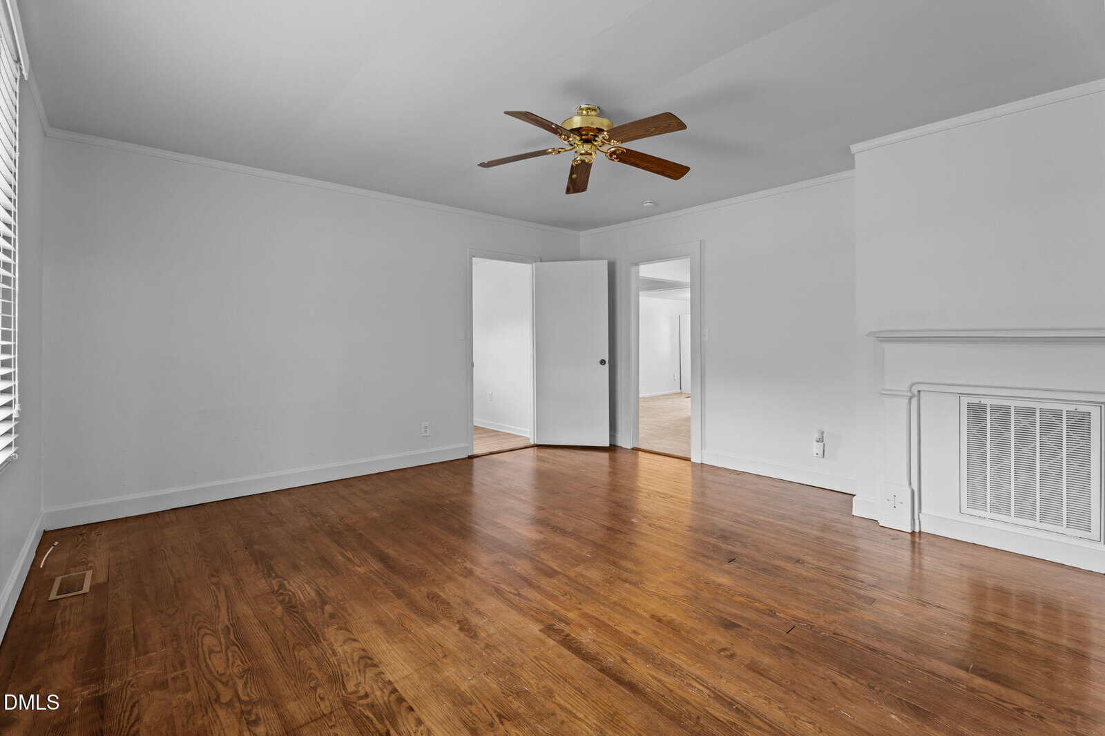 Empty room with hardwood floors, white walls, a ceiling fan with wooden blades, a white door leading to another room, and a vented wall panel near the corner. Natural light enters from the left through window blinds.