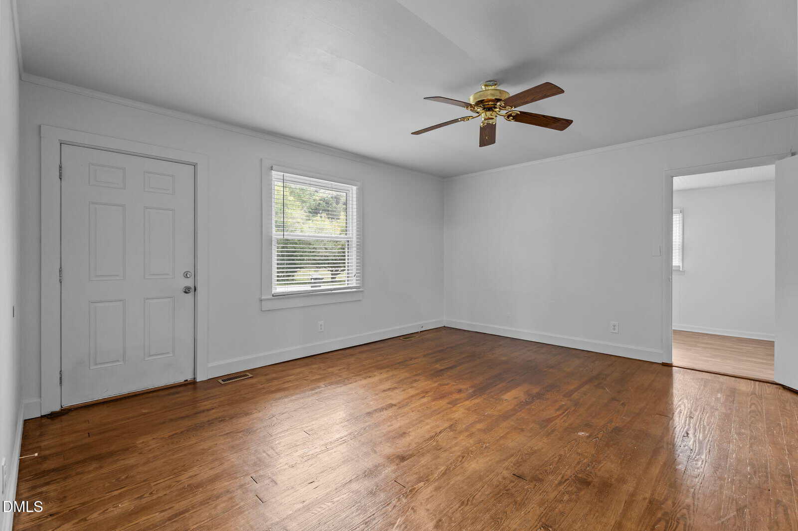 Empty room with white walls, a wooden floor, a ceiling fan, a window with blinds, a closed door, and an open doorway leading to another area. The room appears well-lit with natural light.
