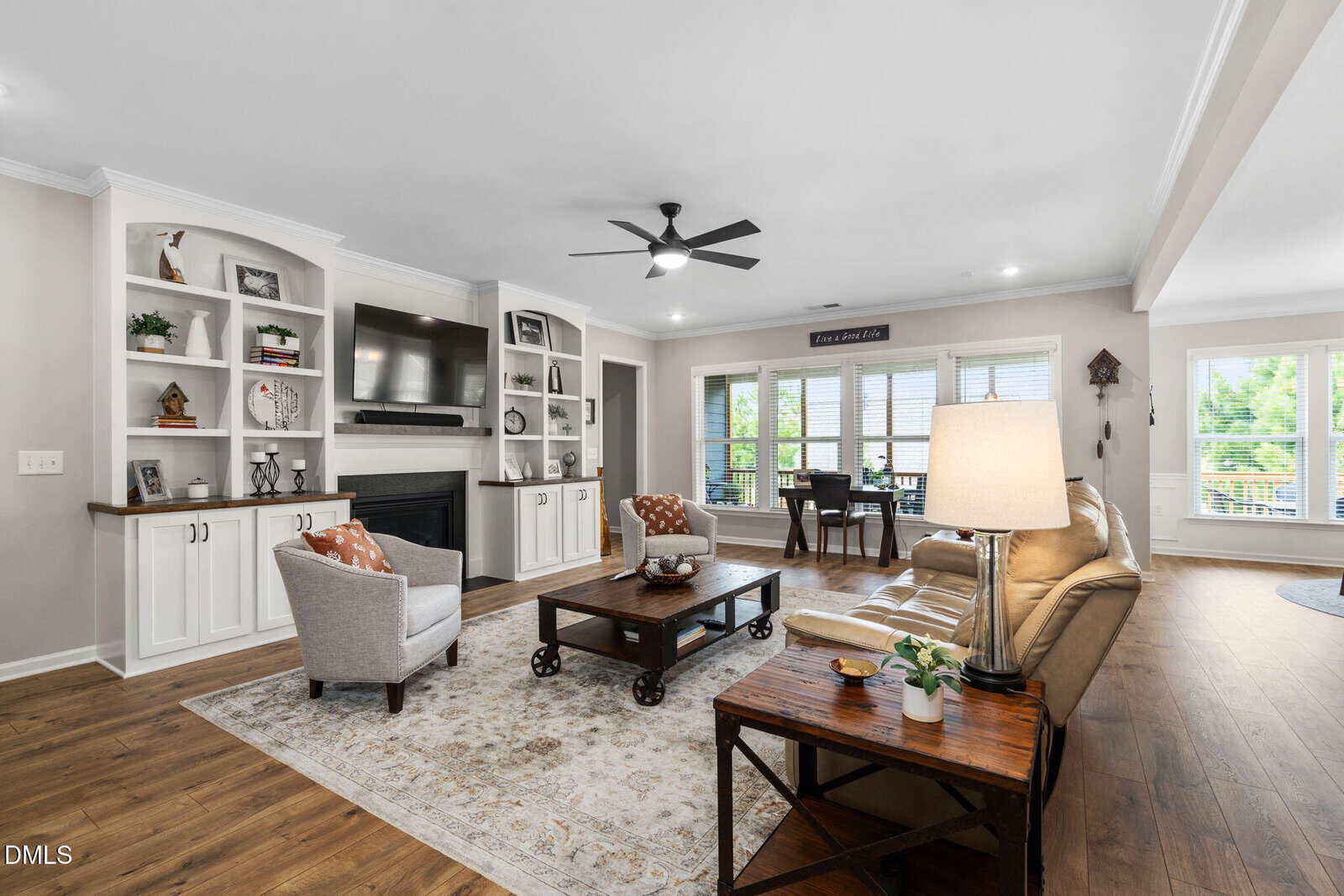 A modern living room with a beige sectional, armchair, coffee table, TV above a fireplace, built-in shelves, wooden floors, and large windows letting in natural light.