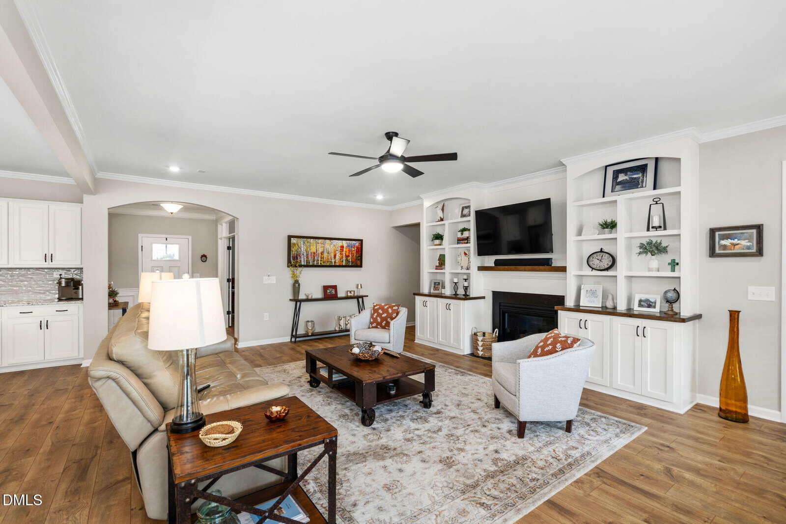 Bright living room with white built-in shelves, a fireplace, TV, beige sofa, two gray chairs, coffee tables, ceiling fan, wood floor, and neutral decor. Large artwork hangs on the far wall by the hallway entry.