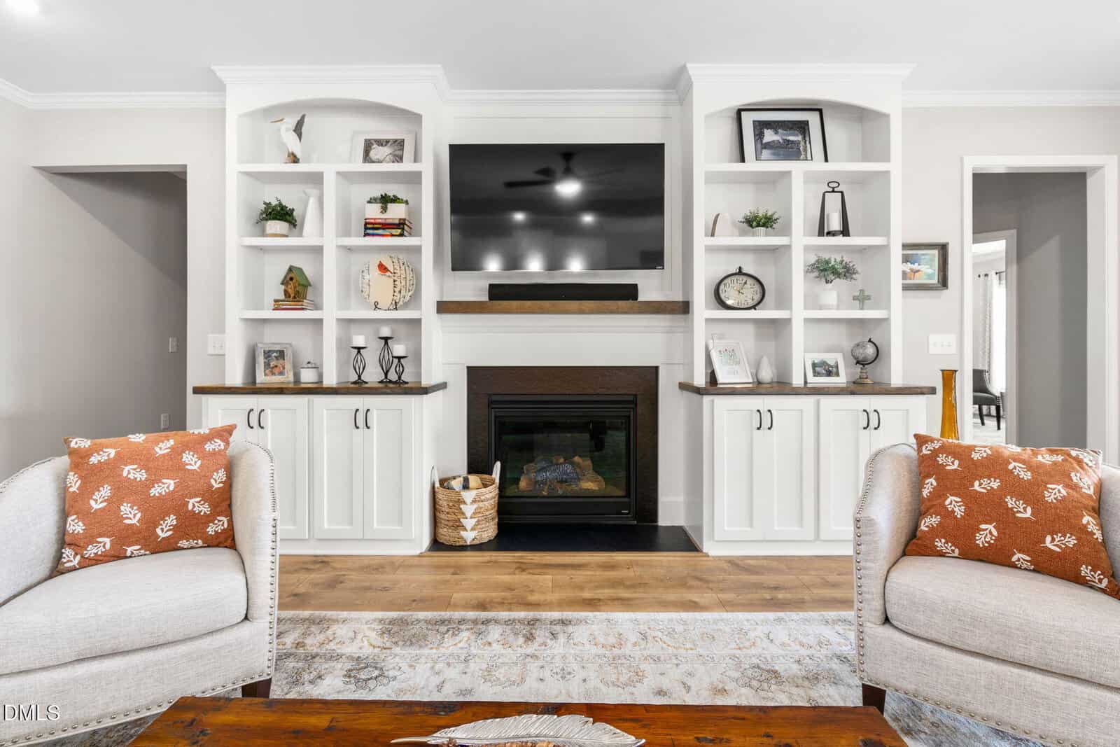 Bright living room with white built-in shelves, a central fireplace, a wall-mounted TV, decorative items, two beige chairs with orange patterned pillows, and a wooden coffee table on a rug.