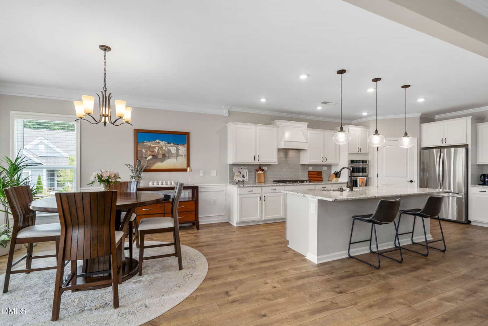 Modern kitchen and dining area featuring white cabinetry, stainless steel appliances, marble island with two barstools, round dining table with chairs, pendant lights, and wood flooring. Large window lets in natural light.
