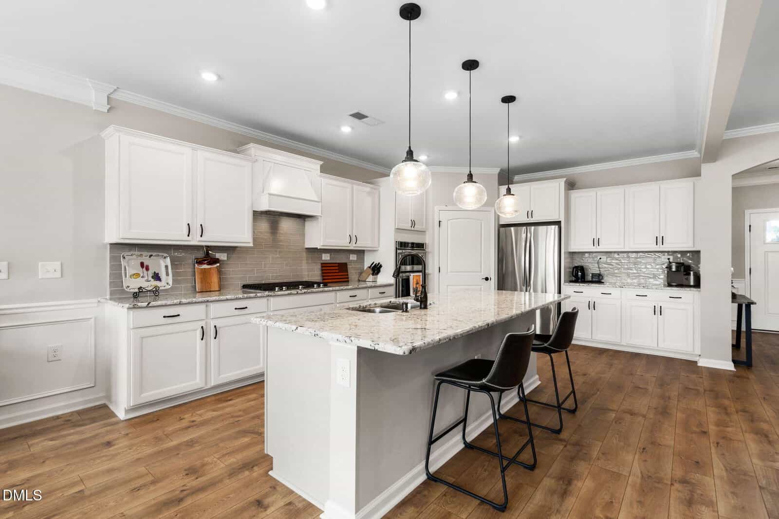 Modern kitchen with white cabinets, stainless steel appliances, a large marble island with a sink, three pendant lights, and two black barstools on wood flooring.