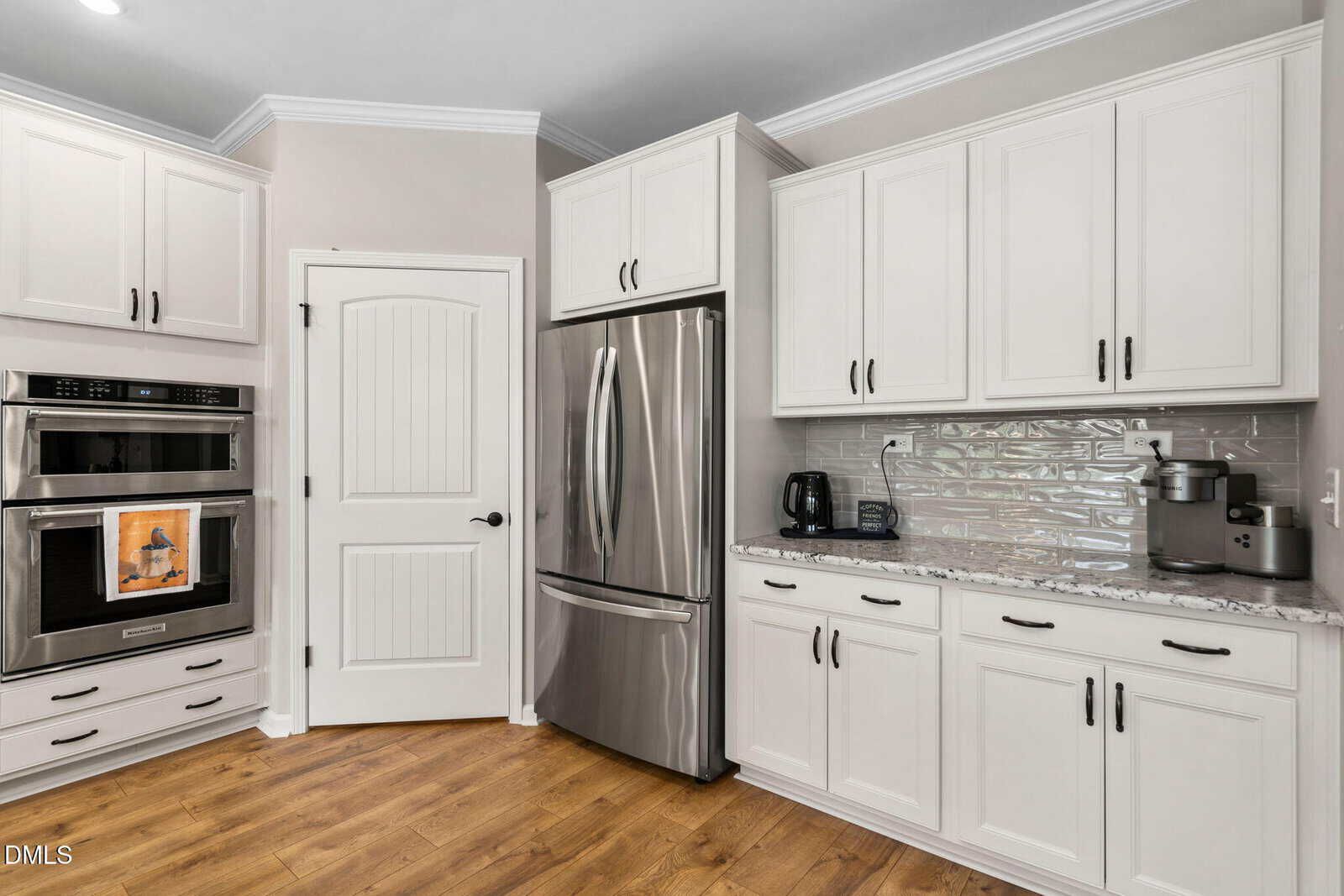Modern kitchen with white cabinets, stainless steel appliances including a refrigerator and double oven, granite countertops, a light tile backsplash, and small appliances on the countertop. Hardwood floor and a white paneled door are visible.