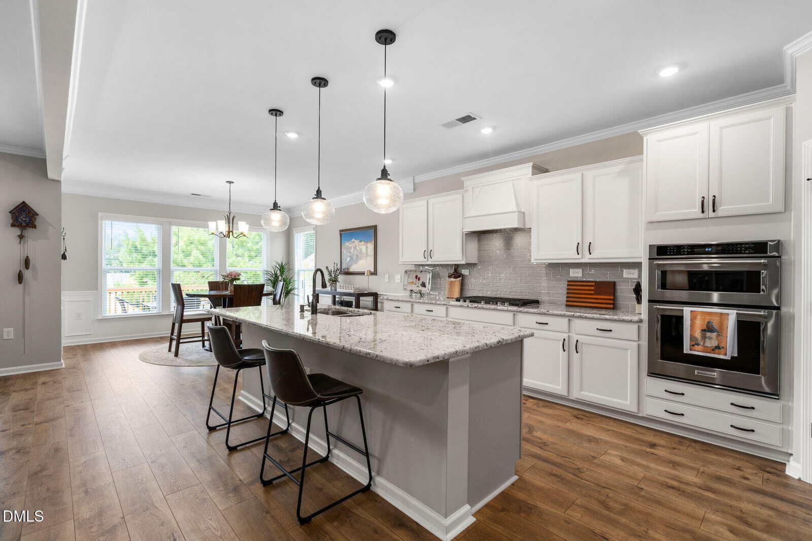 Modern kitchen with white cabinets, a large marble island with sink, three pendant lights, two barstools, and stainless steel appliances; dining area with large windows in the background.