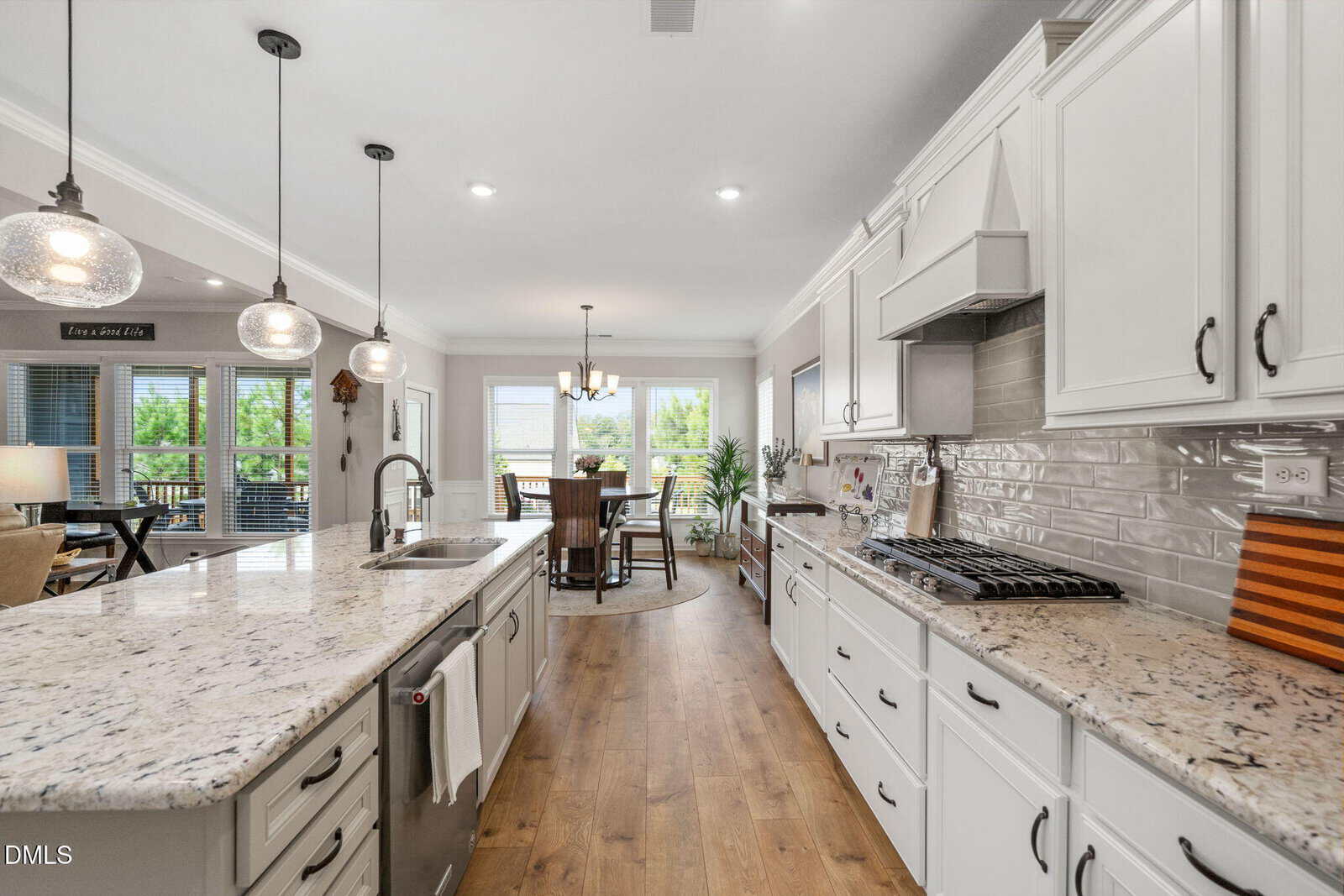 Modern kitchen with white cabinets, granite countertops, island with sink, pendant lights, stainless steel appliances, gas stove, and dining area with large windows overlooking a green outdoor view.