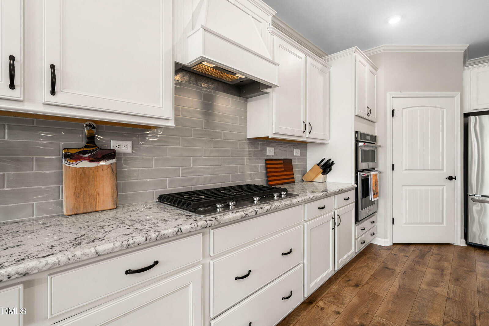 Modern kitchen with white cabinets, marble countertops, gray subway tile backsplash, gas stovetop, stainless steel oven, wood flooring, and various cutting boards and knives on the counter.