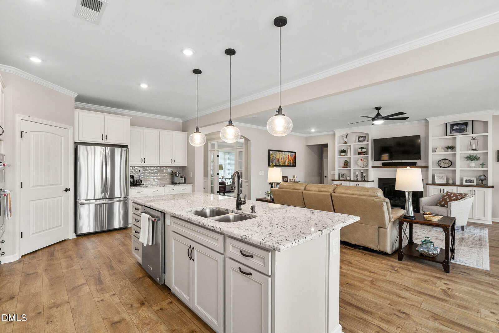 Open-concept kitchen and living area with white cabinets, stainless steel appliances, a large marble island with sink and pendant lights, wood flooring, and a cozy living room with a fireplace and built-in shelves.
