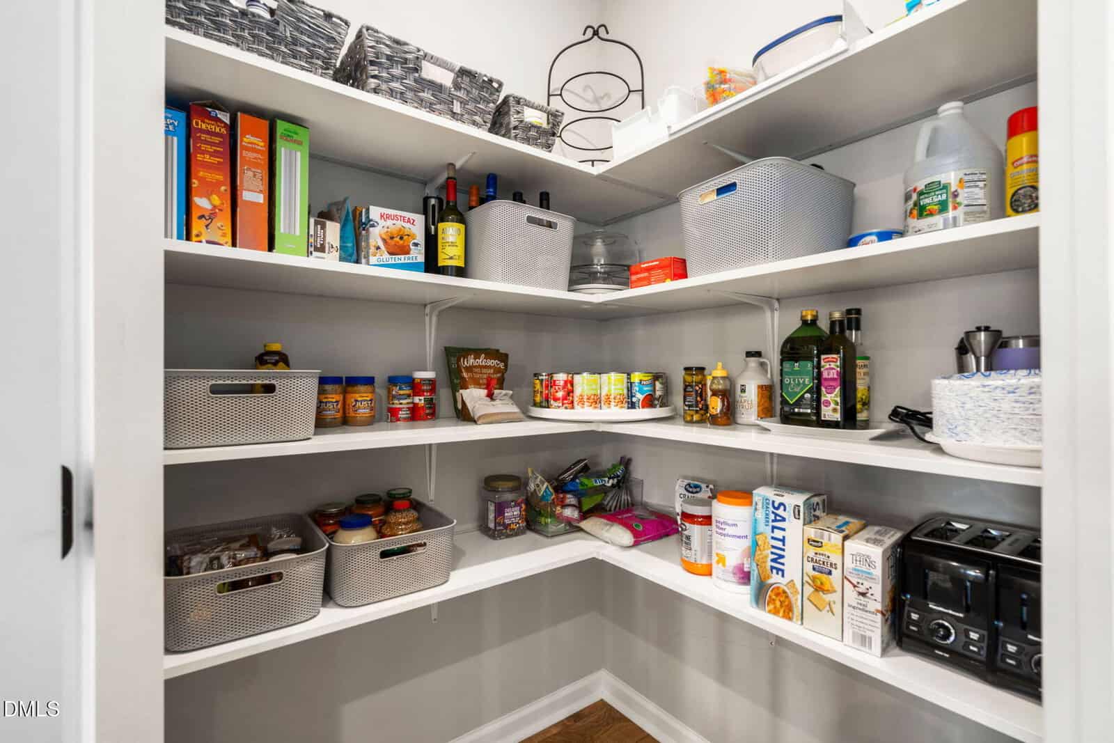 A neatly organized pantry with white shelves holding canned goods, cereal boxes, olive oil, vinegar, snacks, baskets, kitchen appliances, and various food items. The shelves are fully stocked and tidy.