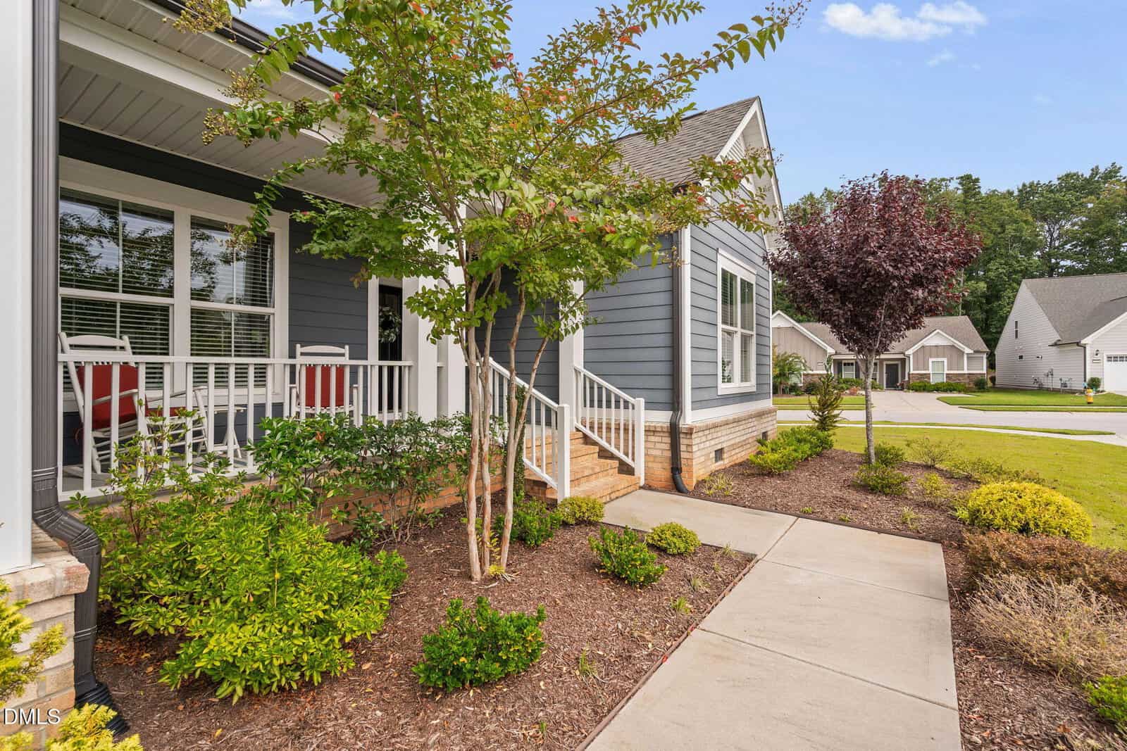 A modern gray house with white trim, a covered front porch with chairs, landscaped yard with shrubs and small trees, and a sidewalk leading to other homes in a suburban neighborhood.