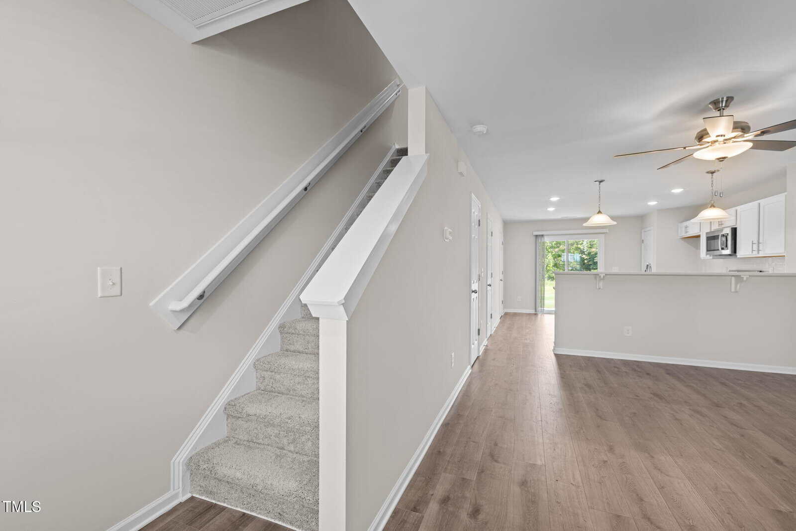 A view of a modern home interior featuring carpeted stairs on the left, wood flooring, neutral-colored walls, a ceiling fan, pendant lights, and a kitchen with a breakfast bar in the background.