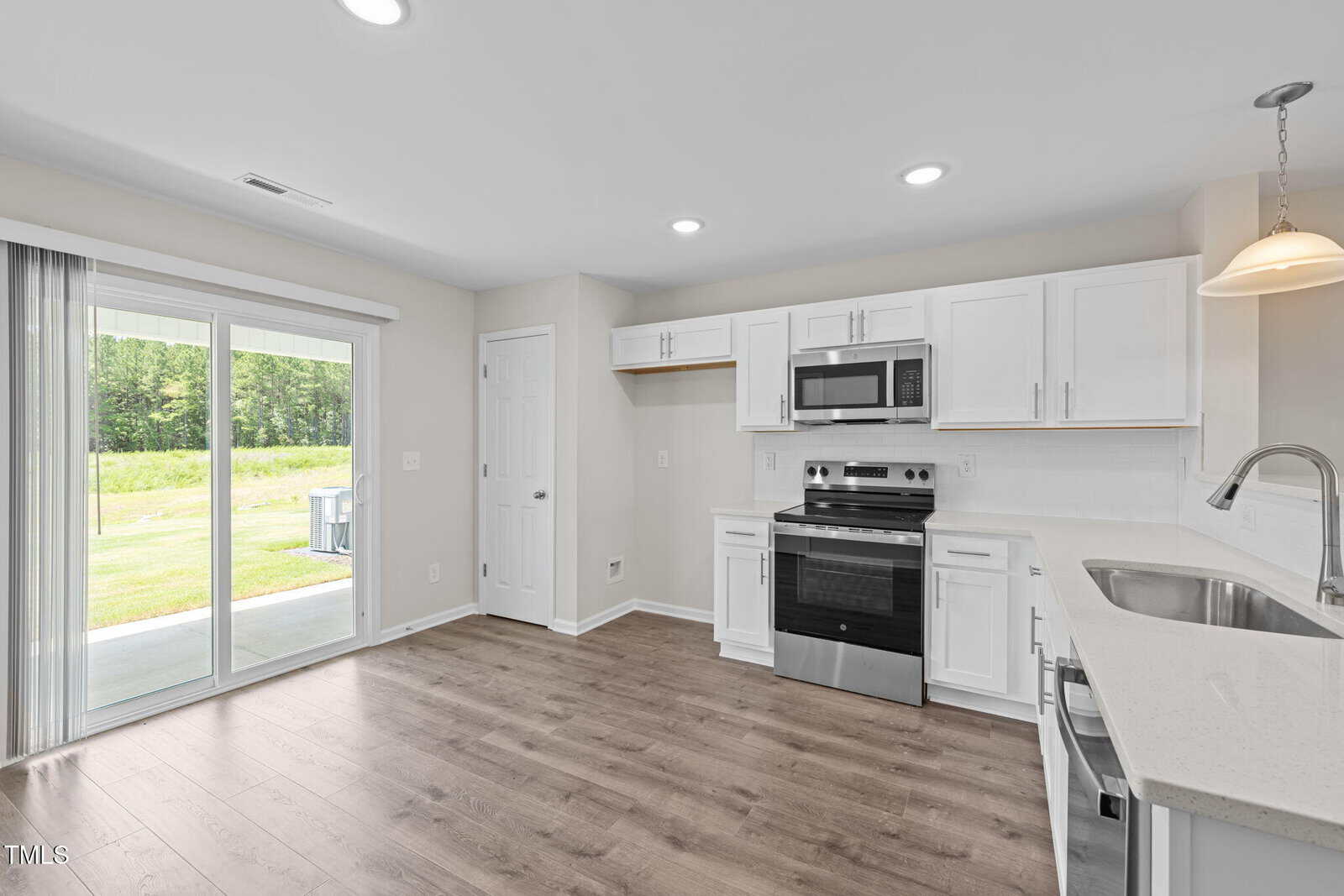 A modern kitchen with white cabinets, stainless steel appliances, light countertops, and wood-style flooring. A sliding glass door opens to a patio and a backyard with grass and trees. A pendant light hangs over the sink.
