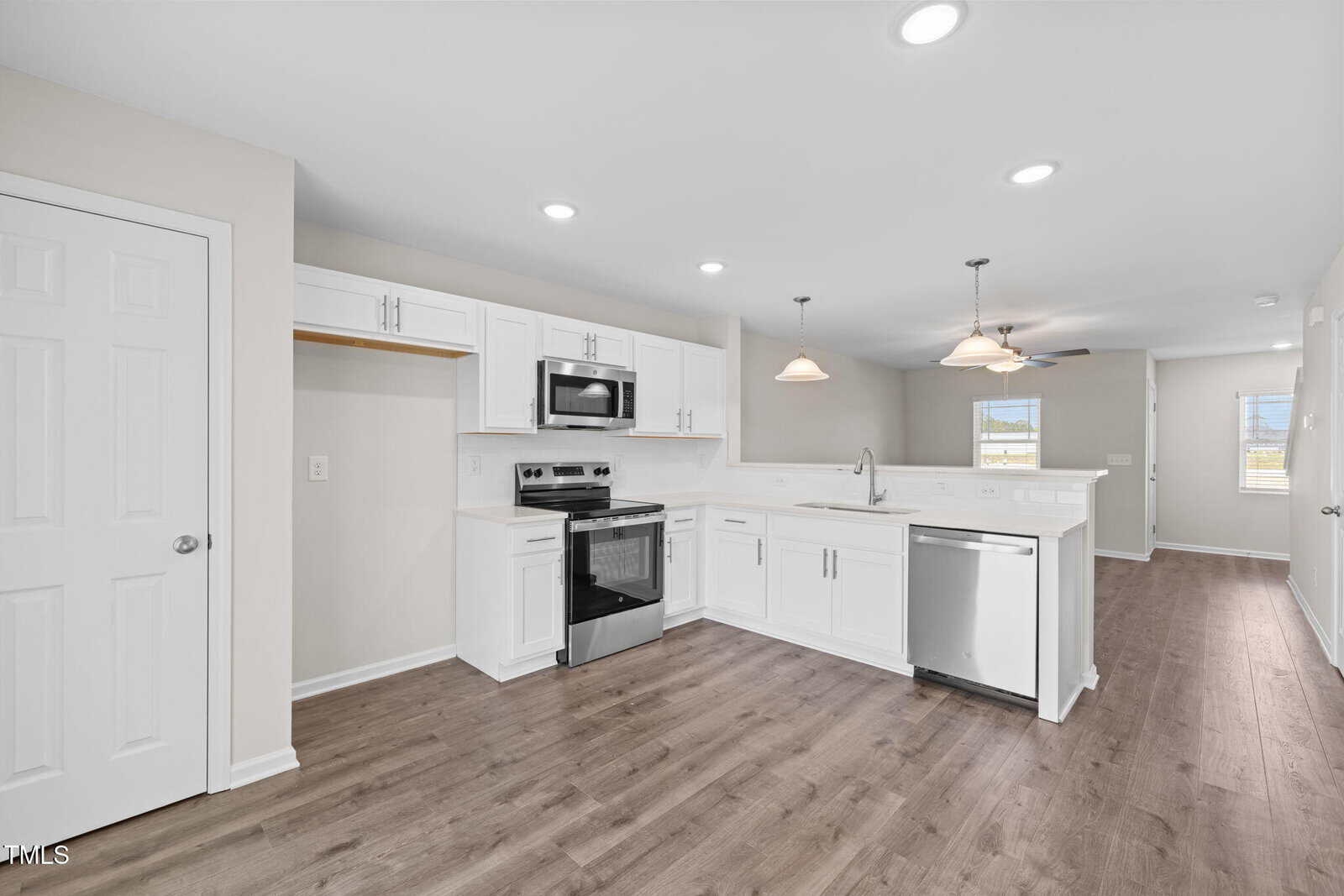 A modern kitchen with white cabinets, stainless steel appliances, wood-look flooring, pendant and recessed lighting, and an open layout extending to a bright living area with windows.