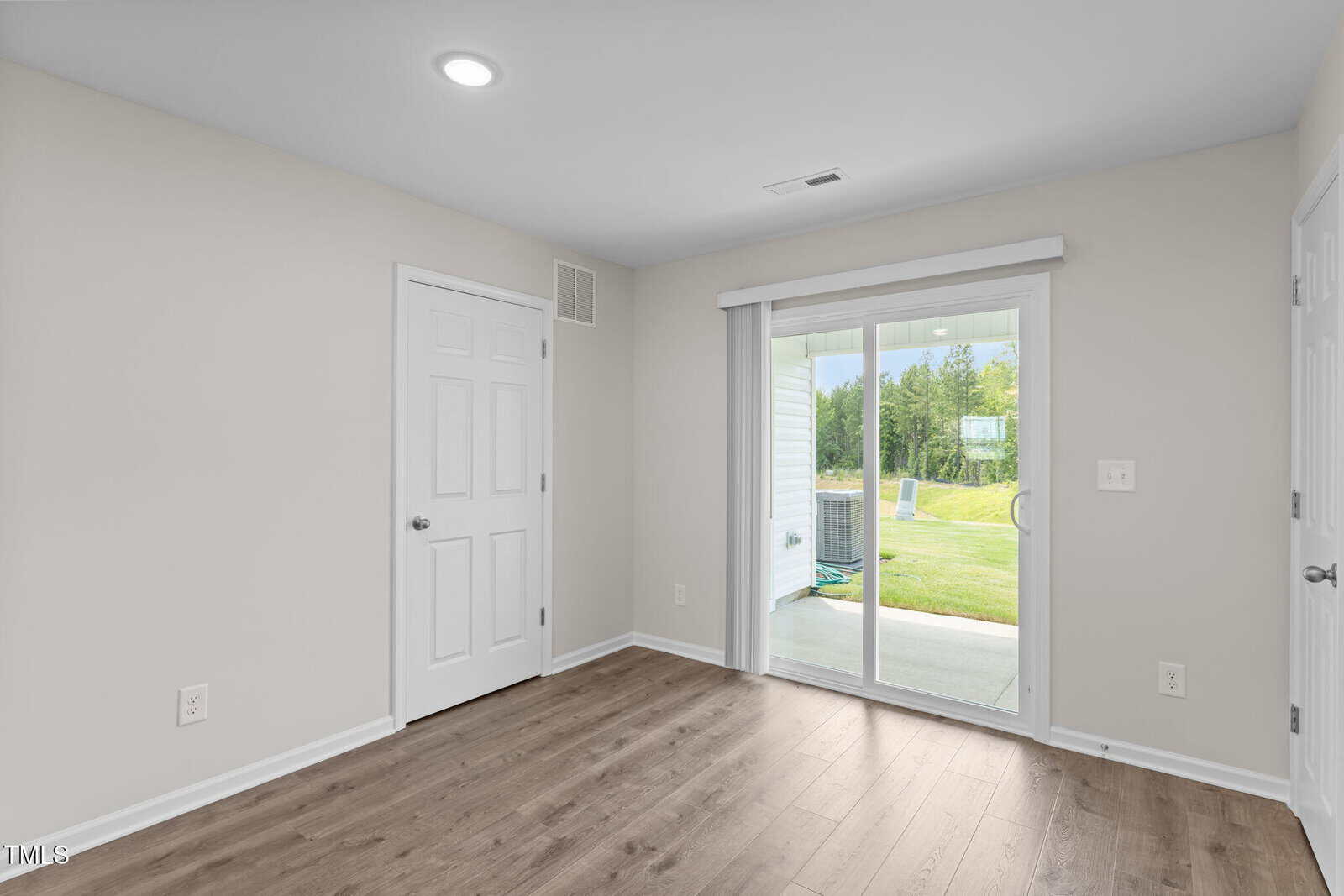 Bright, empty room with light walls, wood flooring, a white door, and a sliding glass door leading to a patio and grassy backyard with trees in the distance.