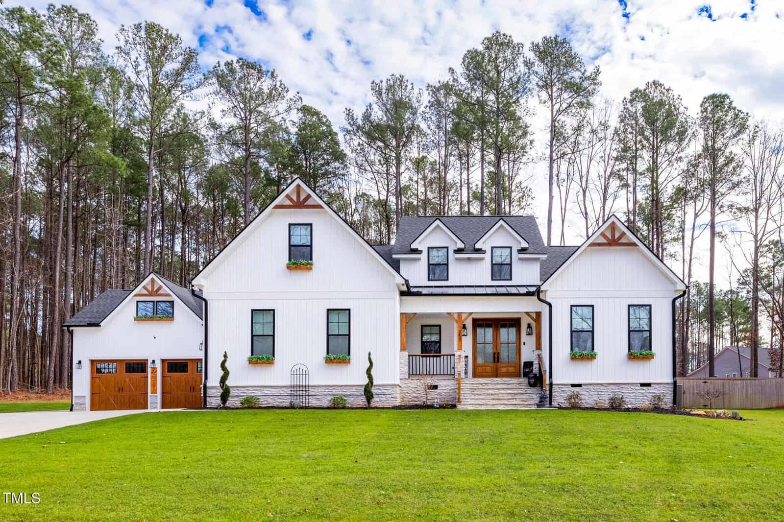 A modern white farmhouse with black trim, wood accents, and a large front porch. The house has a gable roof, dormer windows, a garage with a wooden door, and is surrounded by tall pine trees and a green lawn.
