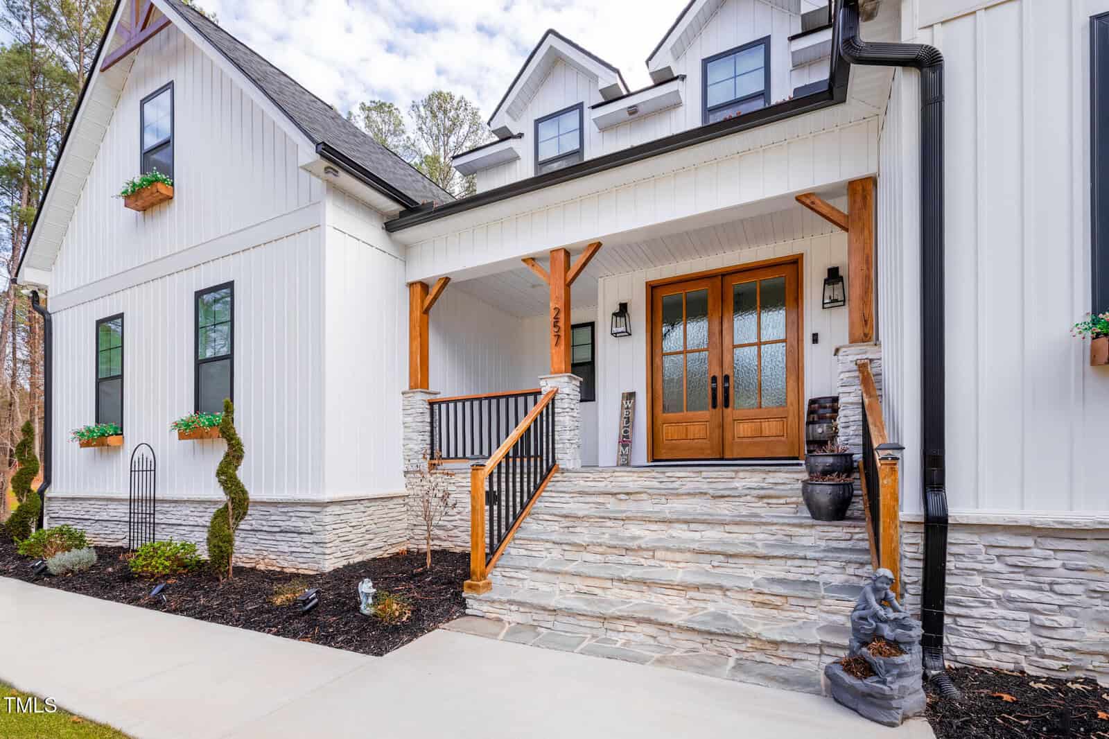 Modern farmhouse with white siding, wood accents, double front doors with glass panels, stone steps, black railings, trimmed bushes, and potted plants; trees and cloudy sky in the background.