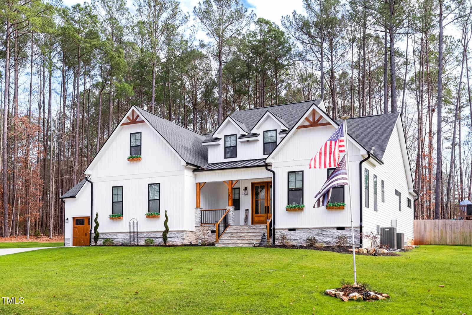 A modern two-story white house with black trim, a covered front porch, and flower boxes under the windows, set on a large green lawn surrounded by tall pine trees, with an American flag in the yard.
