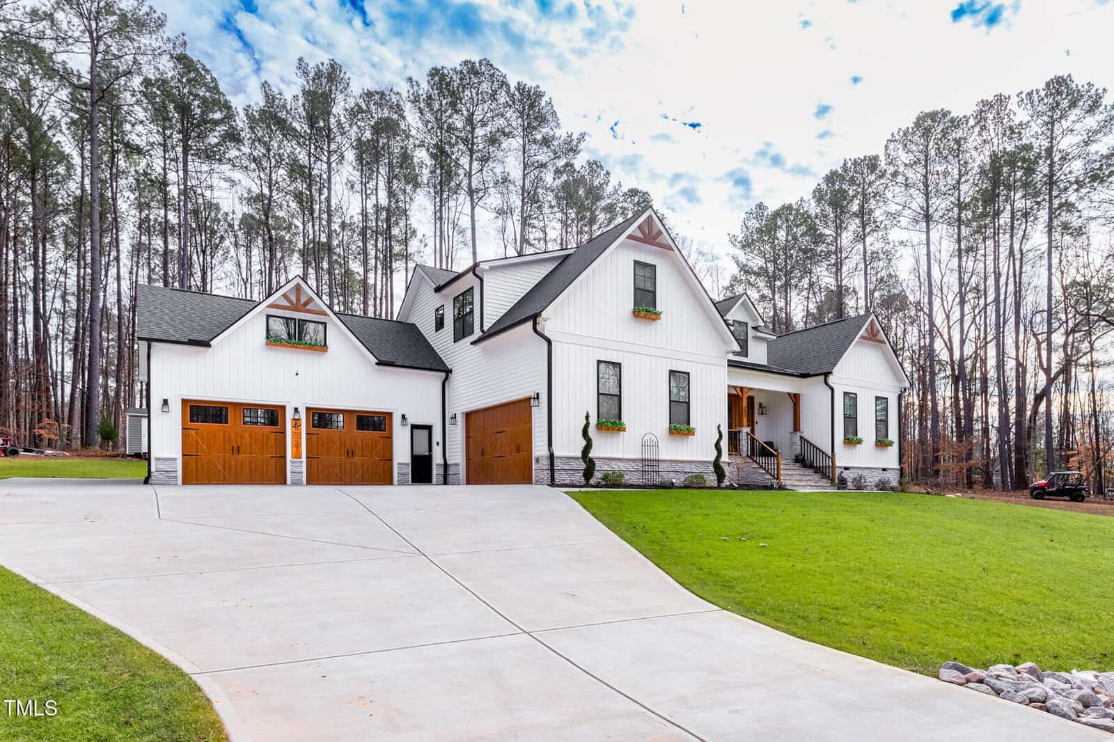 A modern farmhouse-style home with white siding, wood accents, and three garage doors sits on a spacious, green yard with a concrete driveway. Tall pine trees surround the house and flower boxes decorate the windows.