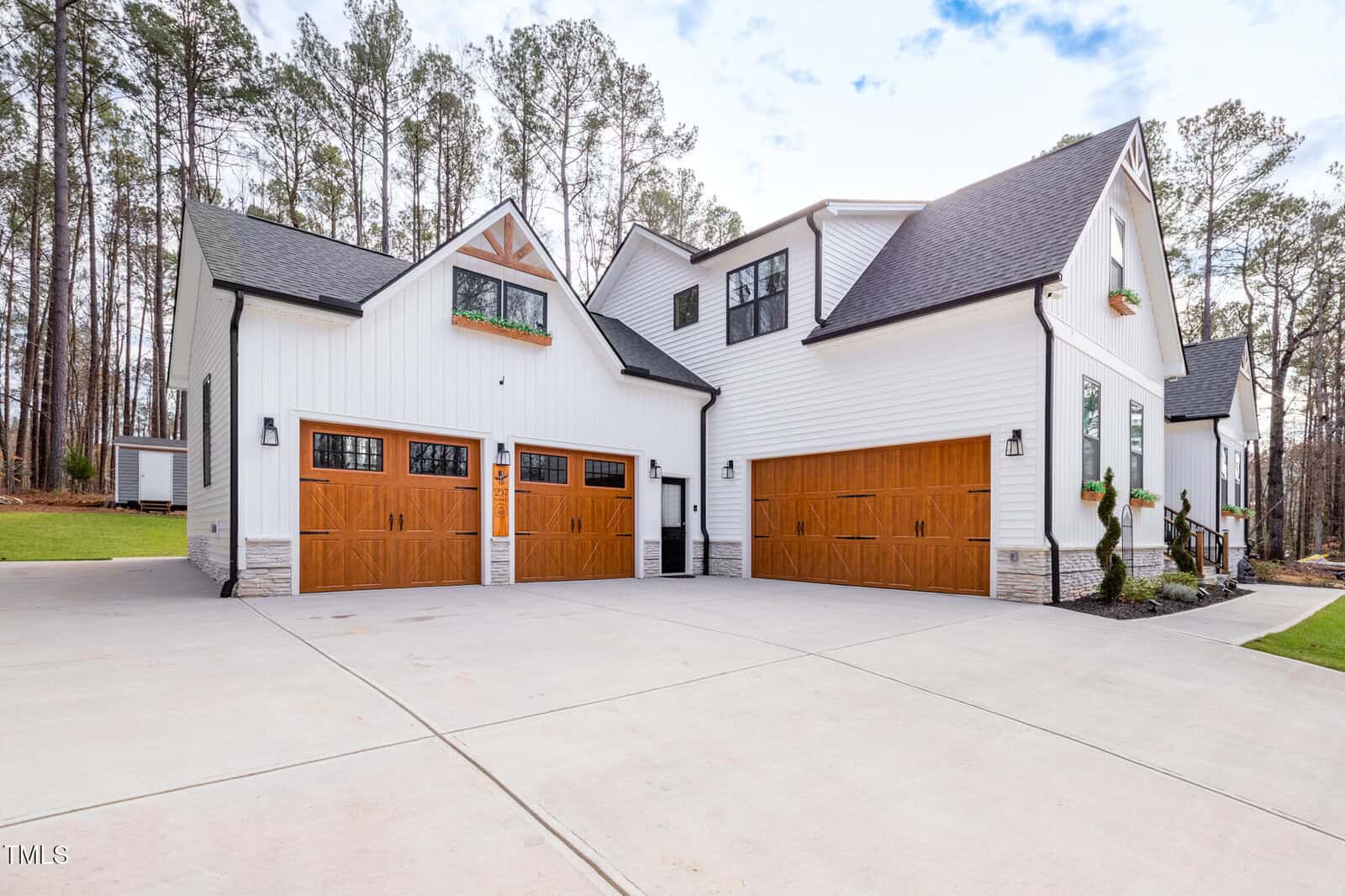 Modern white house with black trim, featuring three wooden garage doors and large windows, surrounded by tall trees. The driveway is wide and paved, and there are window boxes with greenery on the upper windows.